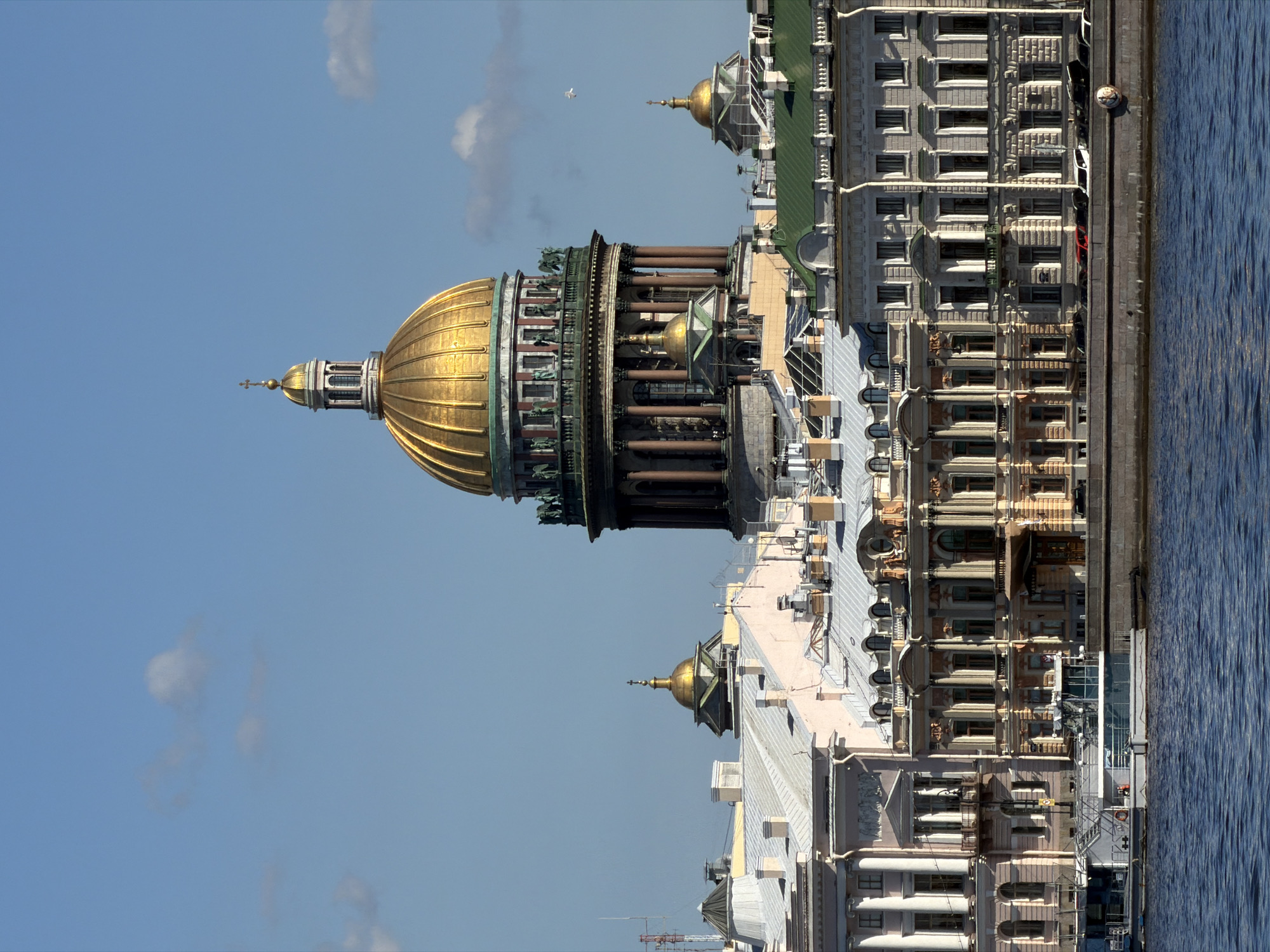 This image depicts a grand cityscape in Saint Petersburg, Russia, showcasing prominent architectural landmarks along a waterway under a bright blue sky.
The central and most dominant feature is a magnificent golden dome, likely belonging to St. Isaac's Cathedral, positioned on the left side of the frame. This large dome is intricately detailed with vertical ribs and topped by a smaller cupola and a cross. Surrounding the base of the main dome are additional architectural elements, including columns and sculptural details.
To the right of the cathedral, a series of stately, multi-story buildings extend horizontally across the middle ground. These structures exhibit classical architectural styles with numerous windows, ornate facades, and some feature smaller, gilded domes or cupolas on their rooftops. The buildings line the edge of a wide body of water, possibly a river or canal, which occupies the lower portion of the image. The water surface shows gentle ripples, reflecting the clear daylight.
The sky above is a vibrant blue, dotted with a few scattered white clouds. A small white bird is visible in flight against the expansive sky. The bright, natural illumination suggests it is daytime. No specific human activity is visible, and there is no discernible text in the image. The scene evokes a sense of historical grandeur and architectural beauty, characteristic of Saint Petersburg.