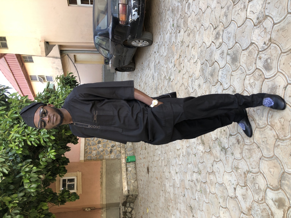 A man wearing glasses, a black traditional outfit, and black shoes with a floral design poses outside. He stands on a patterned stone walkway next to a building with green foliage. A black car is visible in the background, parked next to the building. The man's hand is in his pocket, revealing a gold wristwatch.