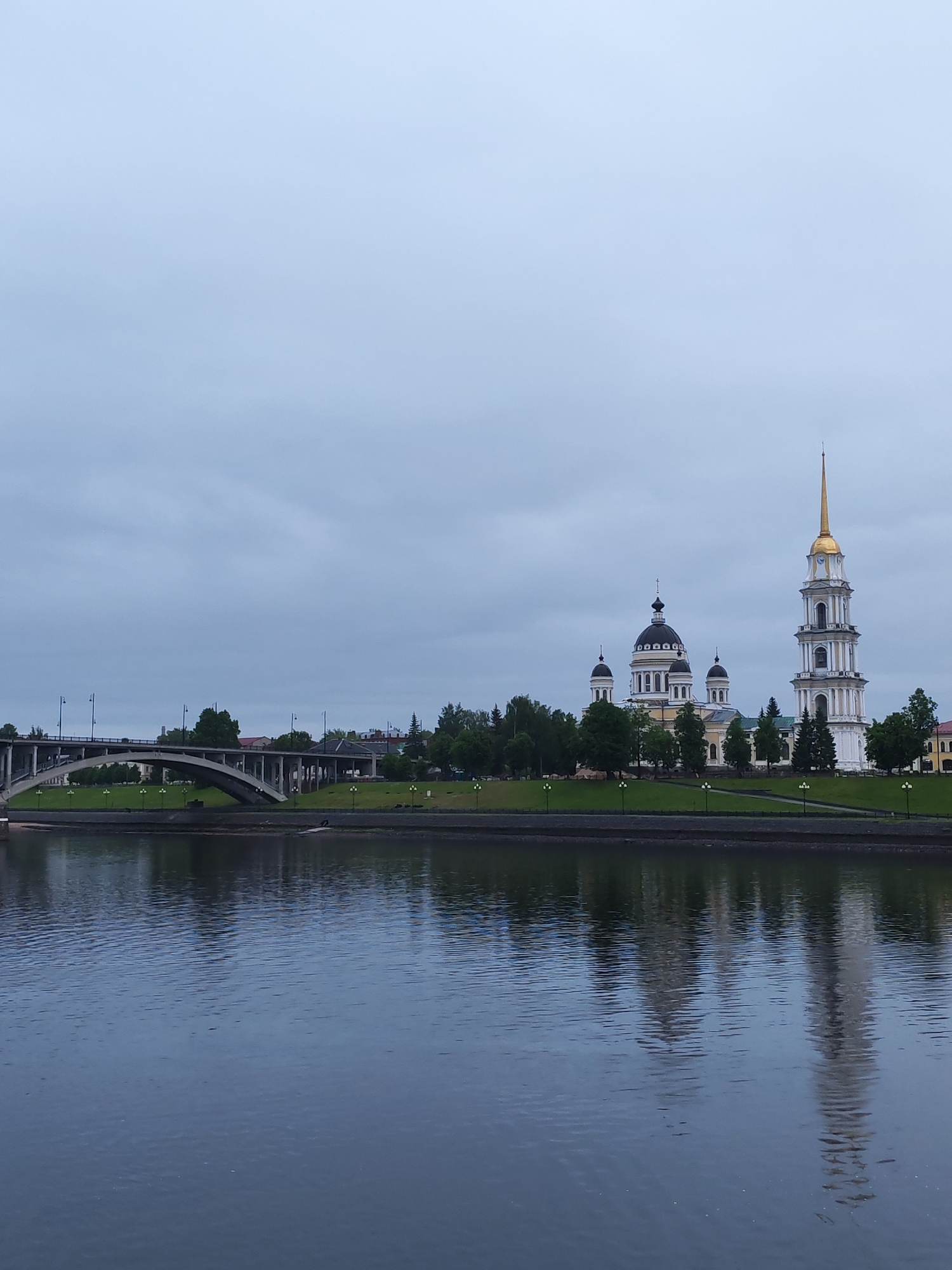 The image depicts a cityscape in Bol'shie Mkhi, Russia, under a cloudy sky. In the midground, there is a river reflecting the buildings and trees on the opposite bank. A bridge spans the river to the left. A prominent Orthodox church with a tall, gilded spire and several black domes is situated on the riverbank amidst green lawns and trees. Lampposts line the riverbank. The river appears still and dark. There are no visible people.