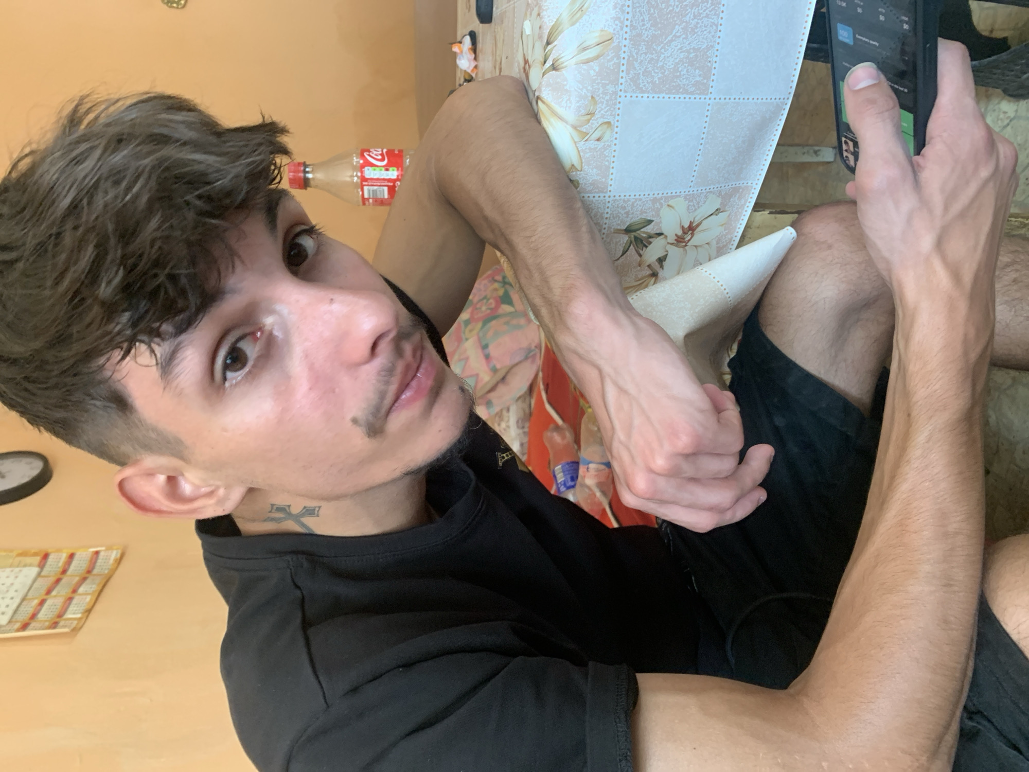 A young man with dark, tousled hair and a faint mustache is shown in a close-up shot, resting his arm on a table covered with a floral tablecloth. He wears a black t-shirt and black shorts. A tattoo of a cross is visible on his neck. He is looking towards the camera, with a Coca-Cola bottle and a calendar in the background. The man is holding a smartphone in his right hand, suggesting he might be taking a selfie or looking at his phone. The scene appears to be indoors, in a casual setting. The estimated location is Mechkarevo, Bulgaria.