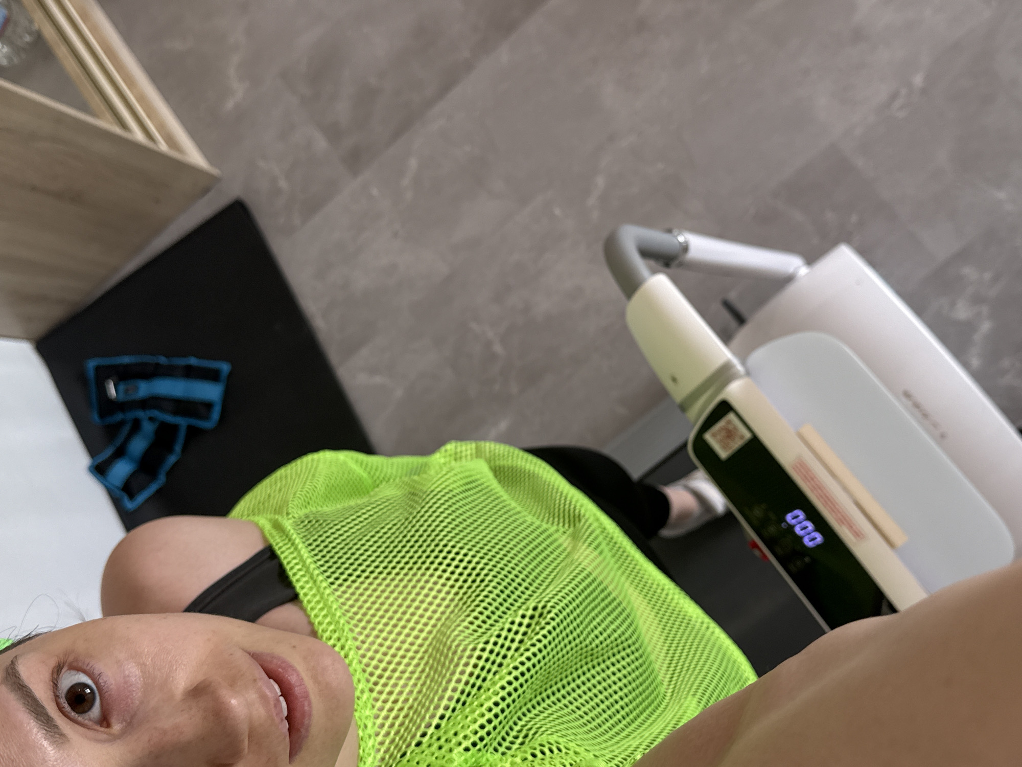 An indoor setting, likely a private residence or home gym, located in Chaikovsky, Russia, is depicted from an elevated, self-portrait perspective.
A female subject occupies the upper left portion of the frame, looking directly into the camera. She has fair skin, a visible brown right eye, and slightly parted lips revealing upper teeth. She is wearing a bright neon green mesh top over a black garment, a matching neon green headband, and black leggings. Her left foot, clad in a white shoe, is positioned on a light grey and white exercise machine.
The exercise machine, possibly a body composition scale, features a black digital display showing "000" in blue digits and a grey handle with a light-colored grip. In the upper right background, two blue and black ankle weights rest on a black exercise mat. Adjacent to the mat, a light wooden cabinet with clear bottles on a shelf is partially visible. The floor throughout the scene is a light grey, possibly tiled or laminated, with a subtle pattern. The lighting is uniform and bright, suggesting an indoor environment.