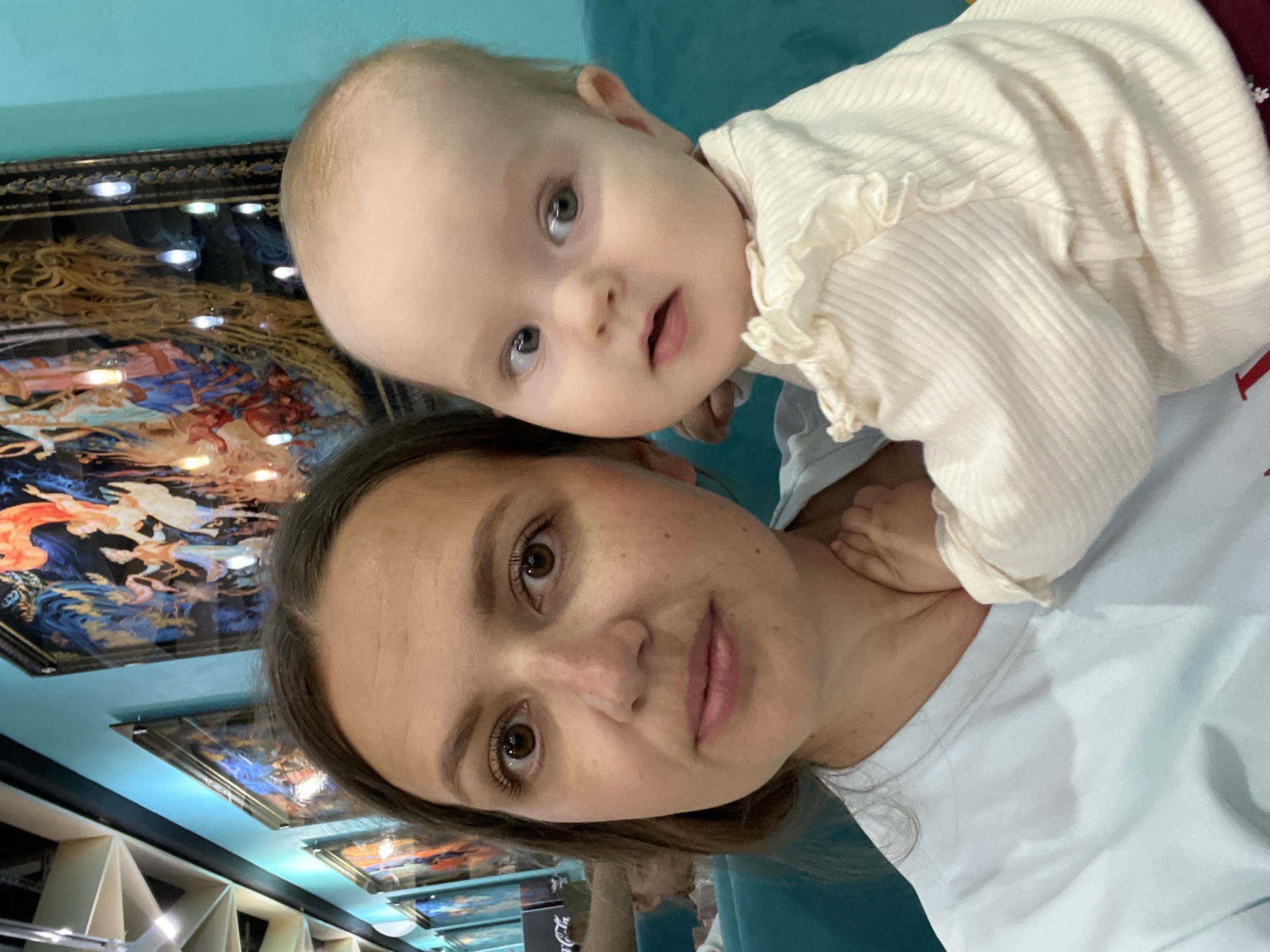 A mother and her baby are posing for a selfie. The mother is holding the baby, who is looking to the side. They are in a restaurant with turquoise walls and paintings on the wall. The mother is wearing a light blue t-shirt and the baby is wearing a cream-colored sweater with ruffles on the collar and sleeves. The background shows a row of paintings on the turquoise wall and a ceiling with wooden beams. A portion of a Coca-Cola sign is visible in the background.