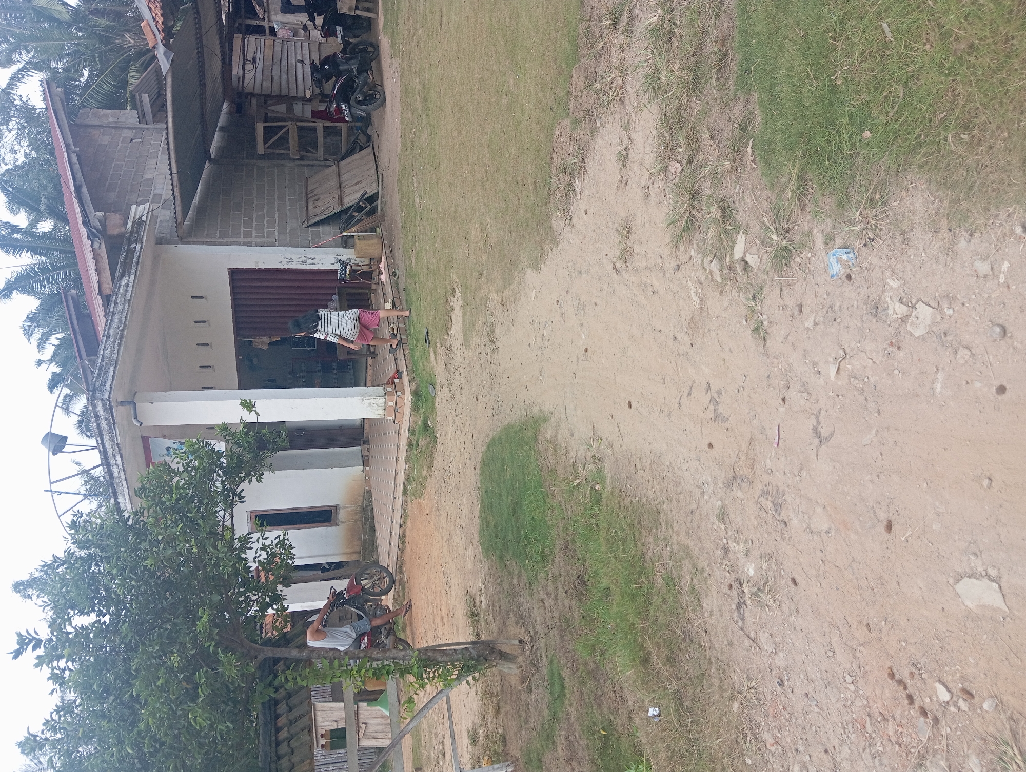 The image depicts a residential scene in Rejosari, Indonesia, on an overcast day. In the foreground, a dirt path with sparse grass leads towards a simple house. The house has a white-painted facade with a large red corrugated metal garage door and a tiled patio area. A man wearing a light blue tank top and dark shorts is seated on a red motorcycle parked on the dirt path to the left of the house. To the right of the house, a young girl with dark hair, wearing a striped shirt and pink shorts, walks away from the camera. Adjacent to the house, a section of the building is constructed with grey cinder blocks, and a corrugated metal roof extends over a covered area. Two dark-colored motorcycles are parked in this covered area. Various wooden structures and debris are visible behind the motorcycles. In the background, palm trees and other greenery suggest a tropical environment. The overall impression is of a rural or semi-urban dwelling with everyday activities taking place.