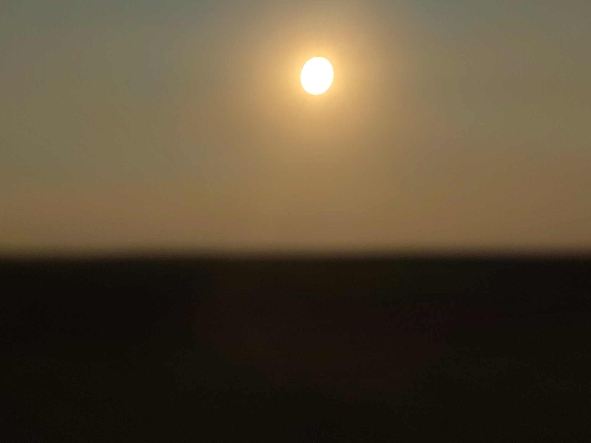 The image depicts a serene sunset scene in Autillo de Campos, Spain. The sun, a bright orb of golden light, hangs suspended in a hazy, gradient sky that transitions from a soft grey at the top to a warm, muted yellow near the horizon. Below the sky, a dark, indistinct band suggests a distant landscape, possibly fields or rolling hills, cloaked in the fading light. The overall impression is one of tranquility and vastness, with the natural elements dominating the frame.