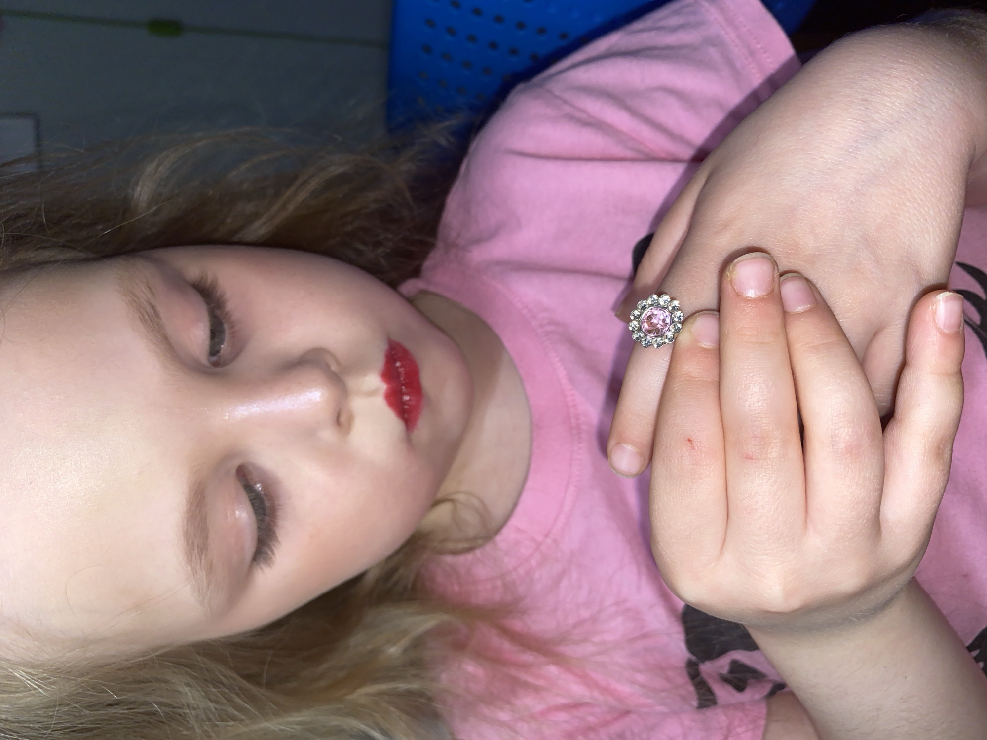 A close-up, high-angle portrait captures a young child lying down, likely indoors. The child has light blonde hair and fair skin, and is wearing a pink t-shirt. A notable detail is the bright red lipstick applied to the child's lips, which appears slightly smudged, hinting at playful self-decoration. The child's eyes are partially closed or cast downwards, giving a serene or contemplative expression.
On the child's right hand, prominently displayed, is a decorative ring. The ring features a large, round pink gemstone in the center, encircled by a halo of smaller, clear sparkling stones. The child's fingernails are short and natural. The fingers are gently curled.
The background is softly out of focus, revealing a dark area above the child's head and a textured blue object, possibly a piece of furniture or a container with perforated details, to the side. The lighting is soft and diffused, highlighting the child's features and the ring. The scene evokes a quiet, possibly sleepy or relaxed moment in an indoor setting, potentially in a home in Votkinsk, Russia. There is no visible text in the image.