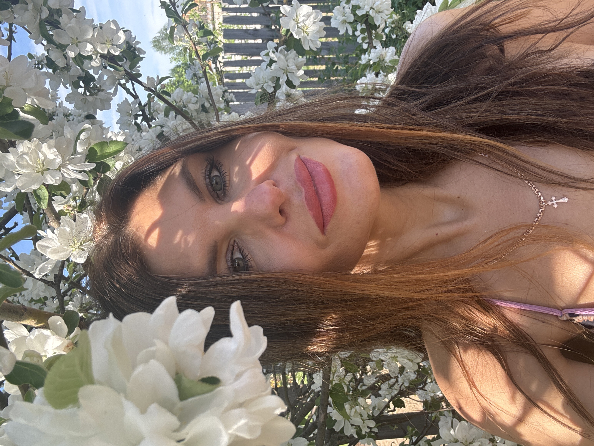 A young woman with brown hair and green eyes poses in front of flowering trees, likely in spring. She is wearing a light pink lipstick and a delicate necklace with a cross pendant. The white blossoms partially obscure a wooden fence in the background. The location in the city of Dubovaya, Russia, is not verifiable based solely on the visual information provided.