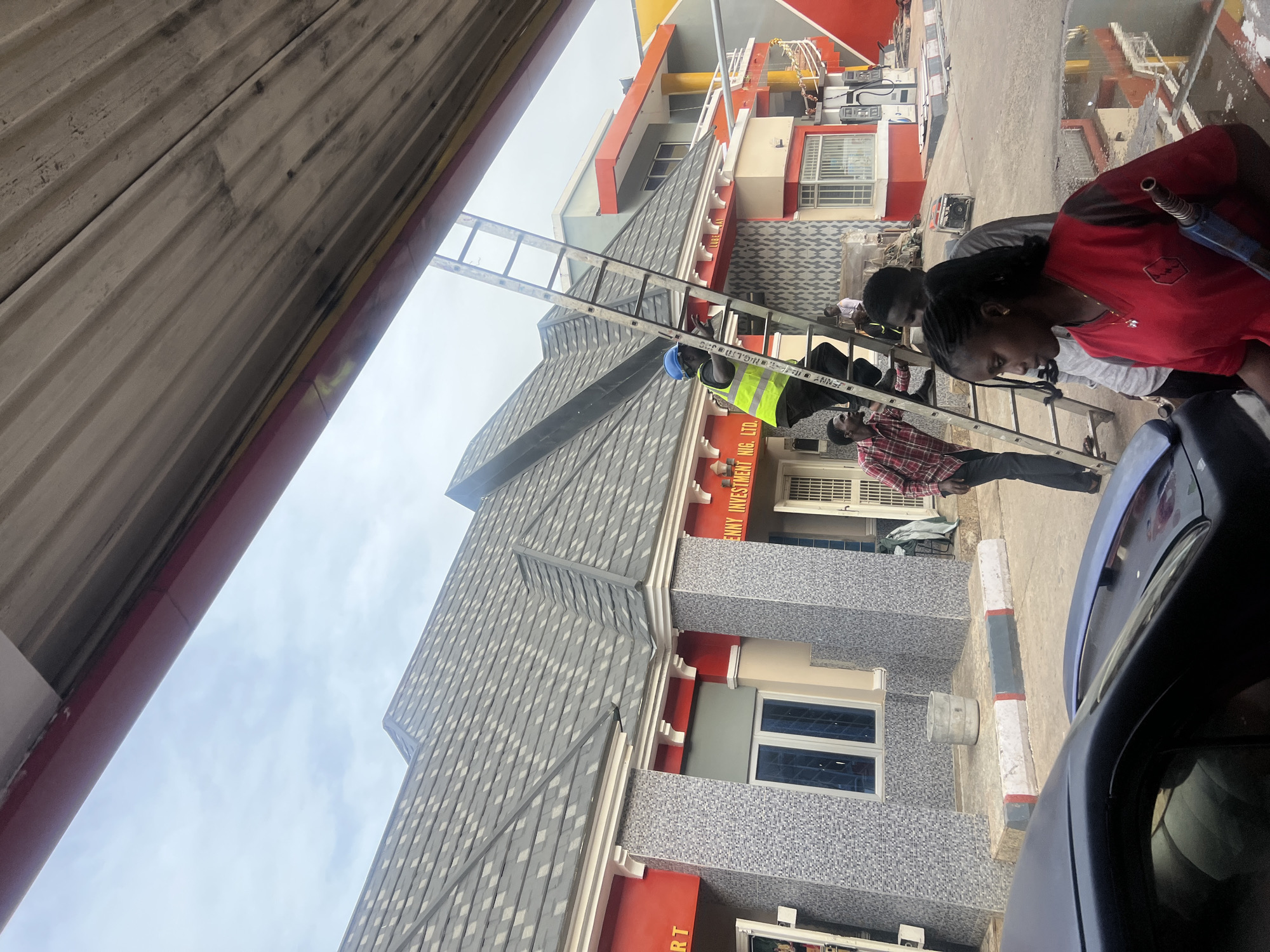 The scene depicts a gas station in Gura Top, Nigeria. A woman in a red shirt is refueling a dark-colored vehicle. A ladder is leaning against a building with gray and red exterior walls, and a man in a yellow safety vest and blue hard hat is standing on it, possibly performing maintenance. Other people are nearby, and the environment suggests a typical urban setting with buildings and street infrastructure.