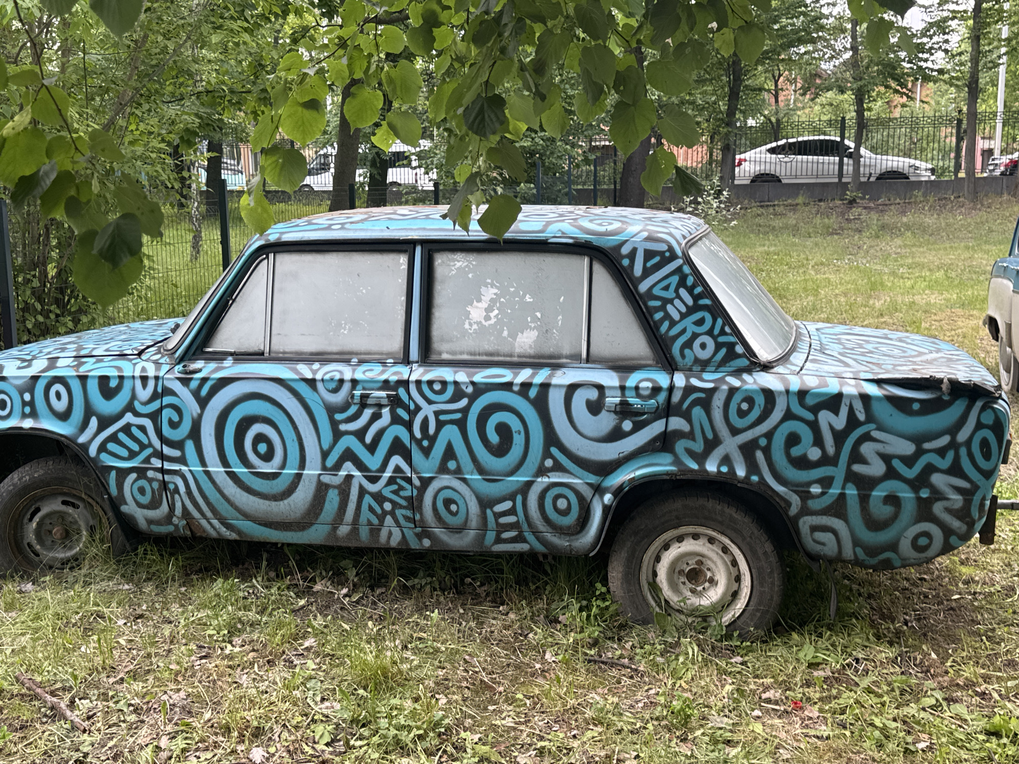 The image shows a vintage blue car, possibly a Lada, parked on a grassy area. The car is heavily decorated with bright blue and black graffiti, featuring swirling patterns and abstract designs. The windows appear to be frosted or covered with a film. In the background, trees, a metal fence, and other vehicles are visible. The overall setting suggests an outdoor urban environment. The presence of a white modern car behind the fence indicates that this is a public space. The graffiti on the car is the most prominent subject in the foreground. This image was captured in Vinnytsia, Ukraine.