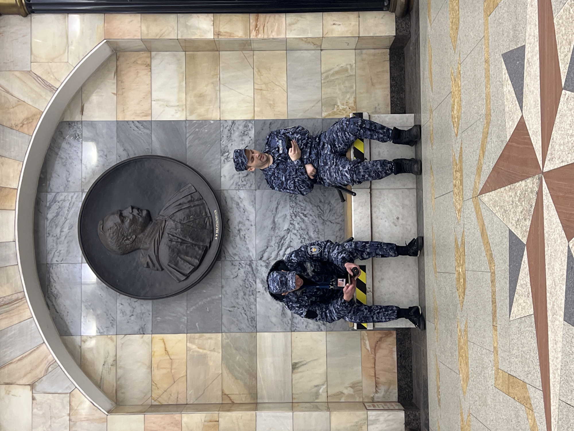 The image captures an overhead view inside a building in Saint Petersburg, Russia. Two uniformed security guards, dressed in camouflage patterned uniforms and boots, are positioned on the floor. One guard is holding and looking at their mobile phone. The other guard is facing towards a large, round bronze bas-relief portrait of a man. The portrait is located within a recessed, arched section of the wall. The floor is composed of marble tiles in varying shades of beige and grey. The tiles have a geometric pattern. The building appears to be well-maintained, and the lighting is bright.