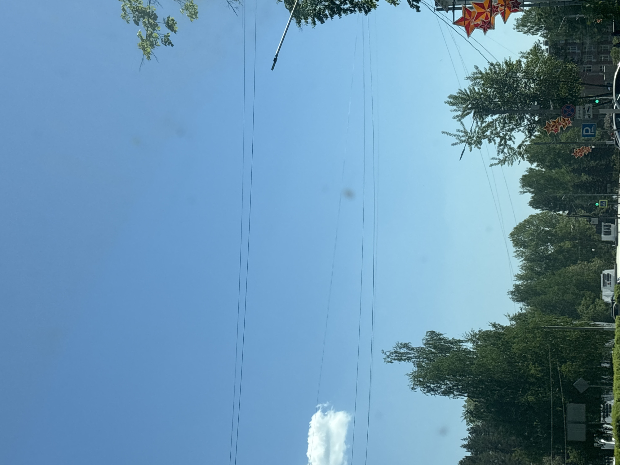 The image shows a street scene in Chaikovsky, Russia. The sky is blue with a few clouds. Power lines are visible overhead. Trees line the street, and decorations, including red stars, are strung up. A parking sign is visible, and various traffic signs and greenery are on the side of the street. Buildings are visible in the background. The image is oriented sideways.