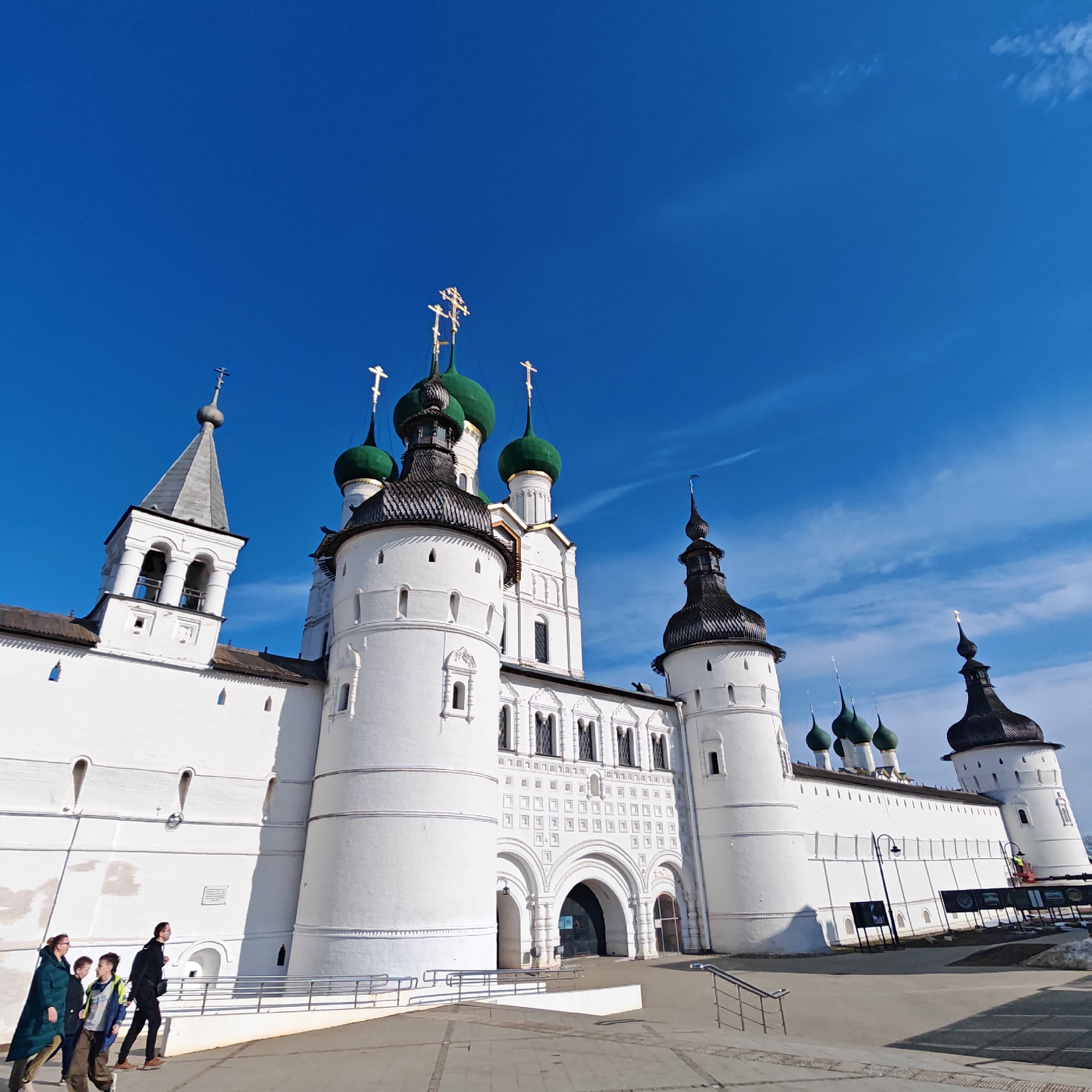 The image depicts the Rostov Kremlin, located in Rostov the Great, Russia, under a clear blue sky. The scene is dominated by a sprawling architectural complex featuring fortified white walls, multiple towers, and churches. Numerous green onion domes, each topped with a golden Orthodox cross, are prominently visible. Several towers have dark, conical or ovoid roofs. In the center, a large white gateway structure with multiple arches and decorative relief patterns serves as an entrance. To the left, a bell tower with arched openings and a grey spired roof rises above the walls.
In the lower left foreground, a group of four individuals walks from left to right. This group consists of one adult female in a dark teal coat, one adult male in a dark jacket and trousers, and two juvenile males in casual jackets. They are walking on a paved pathway. Metal railings are visible along a raised section of the pathway to their right. Bright sunlight casts distinct shadows on the ground and against the white walls, indicating a clear day. In the mid-ground on the right, several large dark display panels on stands are positioned along the base of the wall. Patches of snow are visible on the ground in the far right. The sky is a deep blue with minimal, wispy cloud formations.