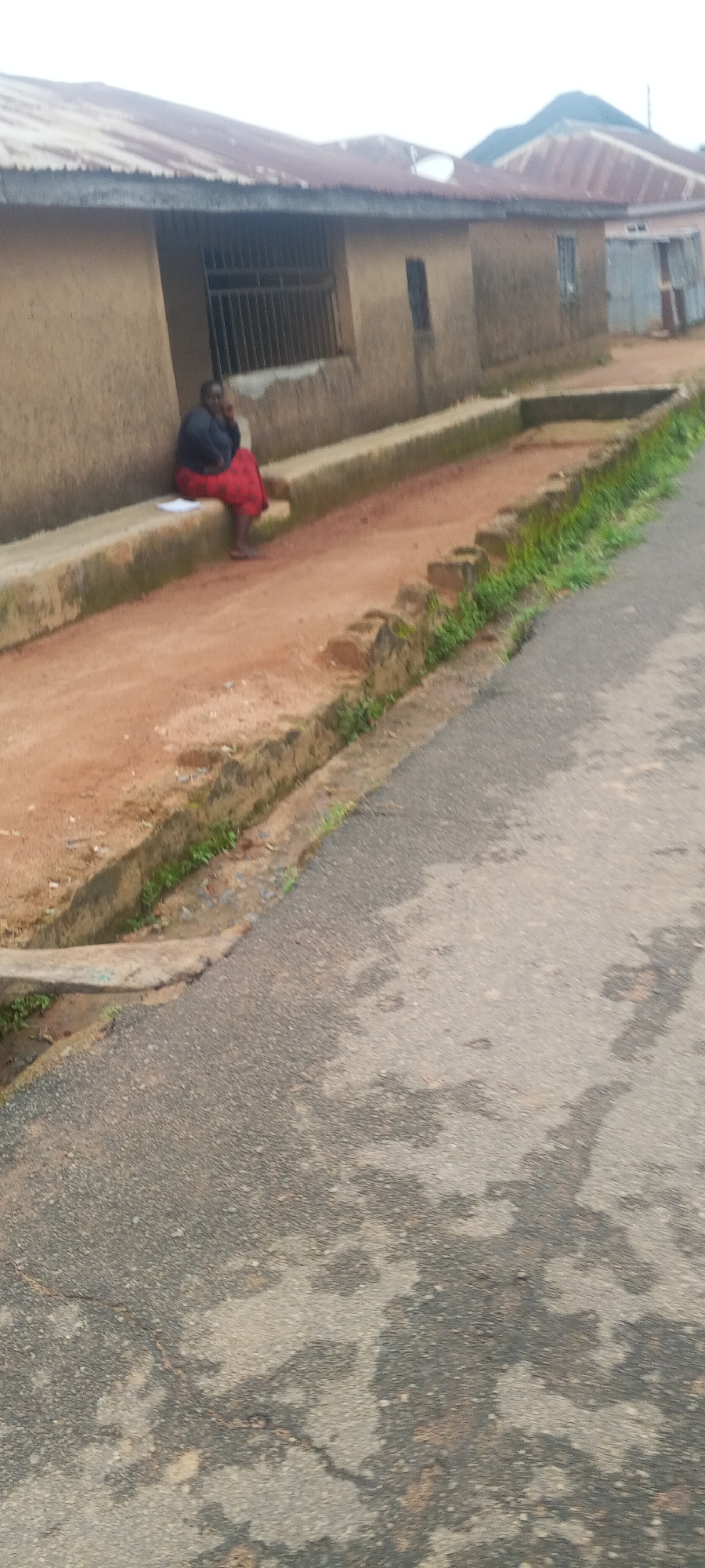 A person is sitting on a concrete ledge outside a mud-brick building with a corrugated metal roof. The person, wearing a dark top and a red and black patterned skirt, is looking down at what appears to be a piece of paper on the ledge in front of them. To the right of the building, a paved road is visible, with a narrow, overgrown drainage ditch running alongside it. The environment appears to be a rural or semi-urban area. The context suggests this scene is located in the city of Bahit, Nigeria.