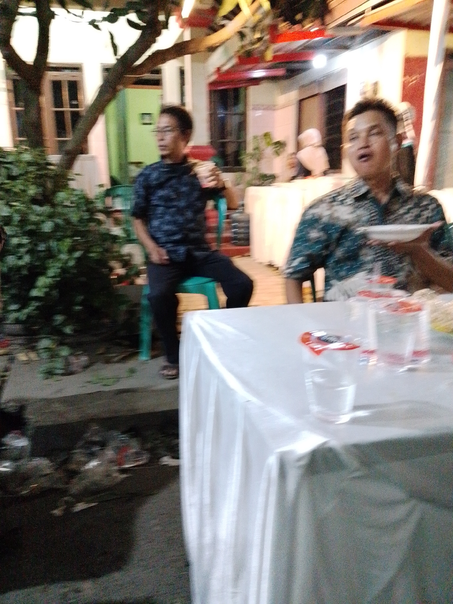 A blurry, nighttime outdoor scene depicts an event in Trangkil, Indonesia. Two men are visible. The man on the left, wearing glasses and a dark patterned shirt, is seated on a green chair. The man on the right, wearing a blue and white patterned shirt, is holding a white plate and appears to be speaking. In the foreground, a table is covered with a white tablecloth, with several plastic cups and bottles on it. A lush green bush is to the left of the seated man, and a tree with dark branches extends from the upper left corner. Buildings with visible windows and lights are in the background.