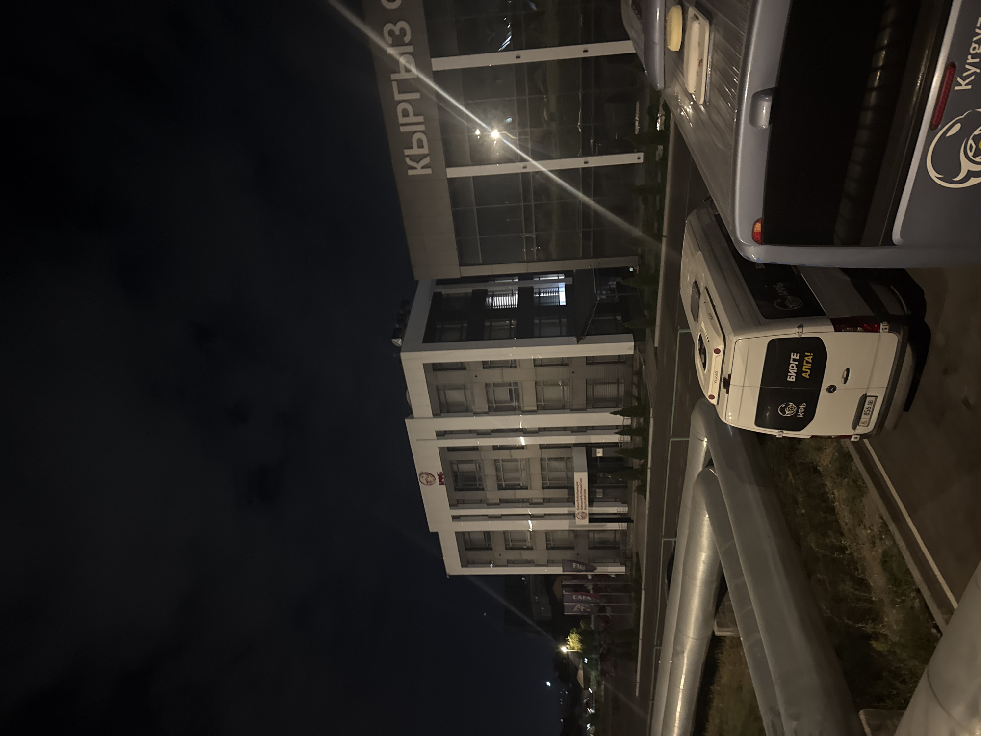 The image shows a modern building at night, with the text "КЫРГЫЗ" visible on the facade. To the left of this building is a smaller, white structure with the number "1995" prominently displayed. In front of the buildings, there are flags planted in the ground. A white van with text in Cyrillic script, including "БИРГЕ АЛГА!", is parked in the foreground, to the right of large, gray pipes that run horizontally across the scene. Another vehicle, possibly a bus, is partially visible on the right edge of the image, also with Cyrillic text on its side. The environment appears to be an urban setting, with streetlights and building lights illuminating the scene against a dark night sky.
