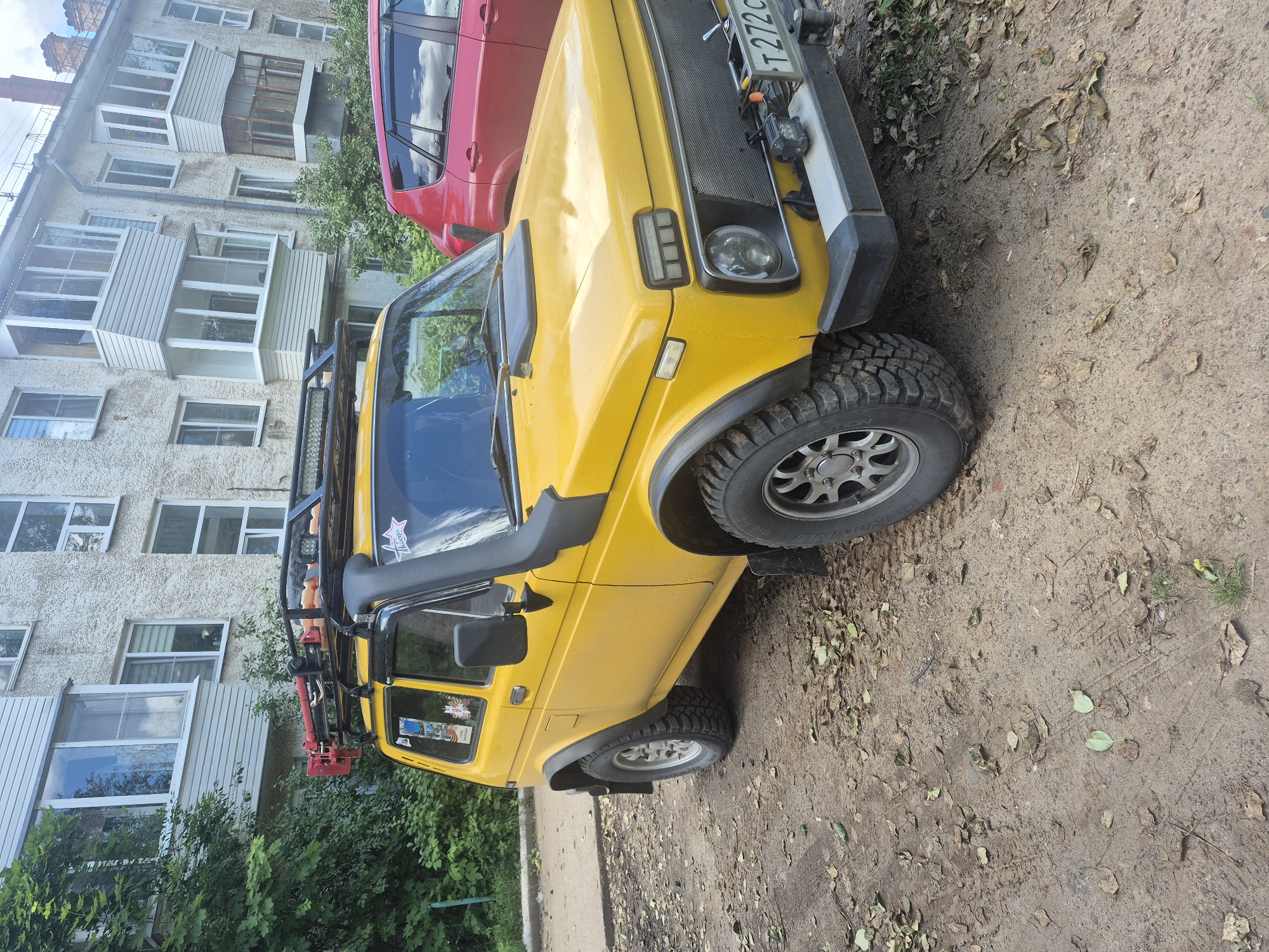 A yellow Niva vehicle is parked on a dirt surface in front of a weathered, light-colored apartment building. The car has a roof rack with what appears to be a Hi-Lift jack attached. A snorkel is fitted to the driver's side A-pillar. The vehicle's front bumper has a license plate that reads "T 272 CS". A red car is partially visible parked to the right of the Niva. Trees and green foliage are present on the left side of the image, and fallen leaves are scattered on the ground. The sky is overcast with clouds visible. The scene is likely in Chaikovsky, Russia, given the appearance of the apartment building and the style of the vehicle.