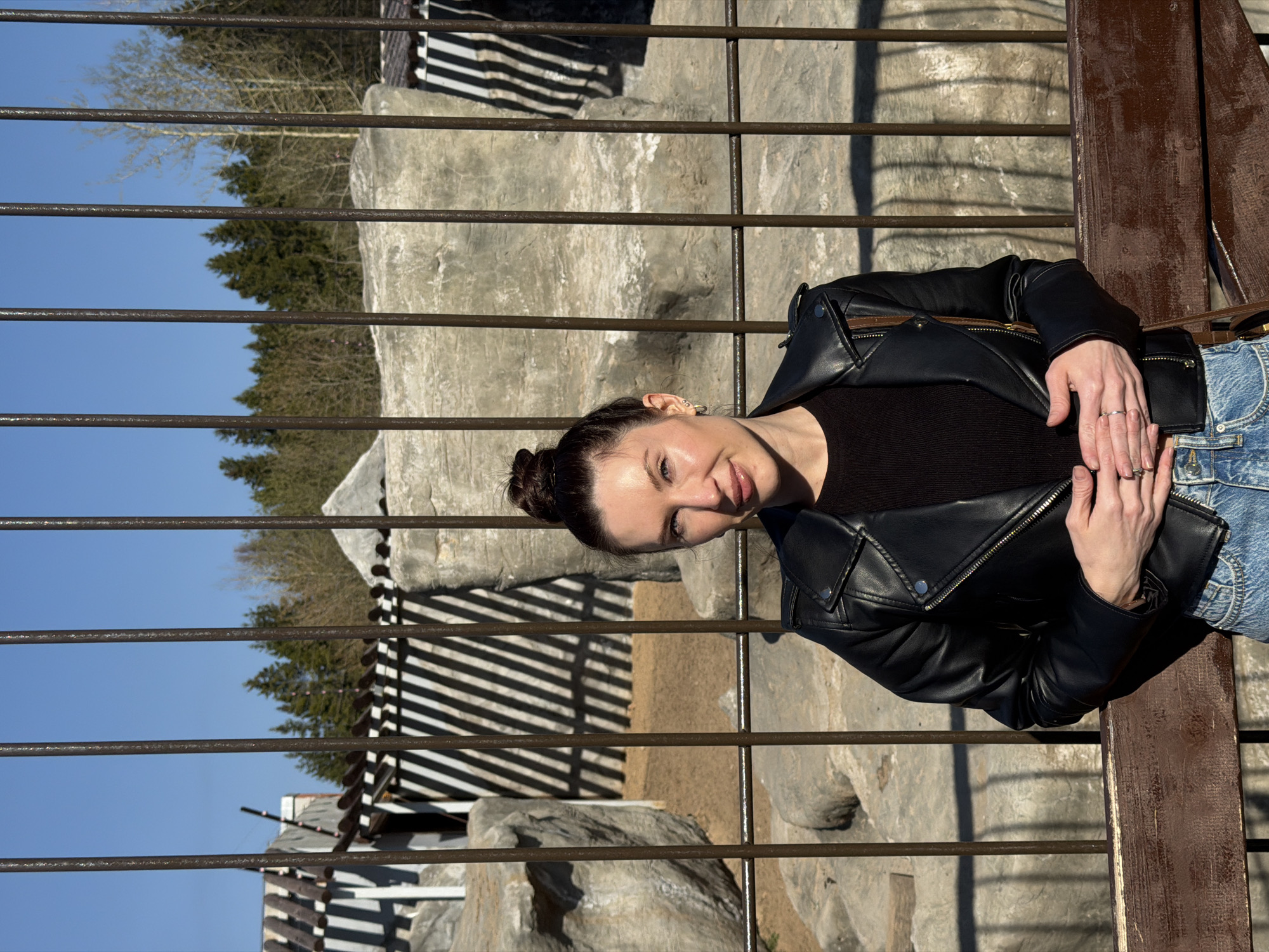 A young woman is sitting on a bench in what appears to be a zoo or animal enclosure. The background has a fence made of metal bars in front of a rocky area. Some buildings are also visible behind the enclosure. The woman is wearing a black leather jacket and blue jeans. She has her hands clasped together in front of her. The scene is likely in Perm, Russia, given the provided context.
