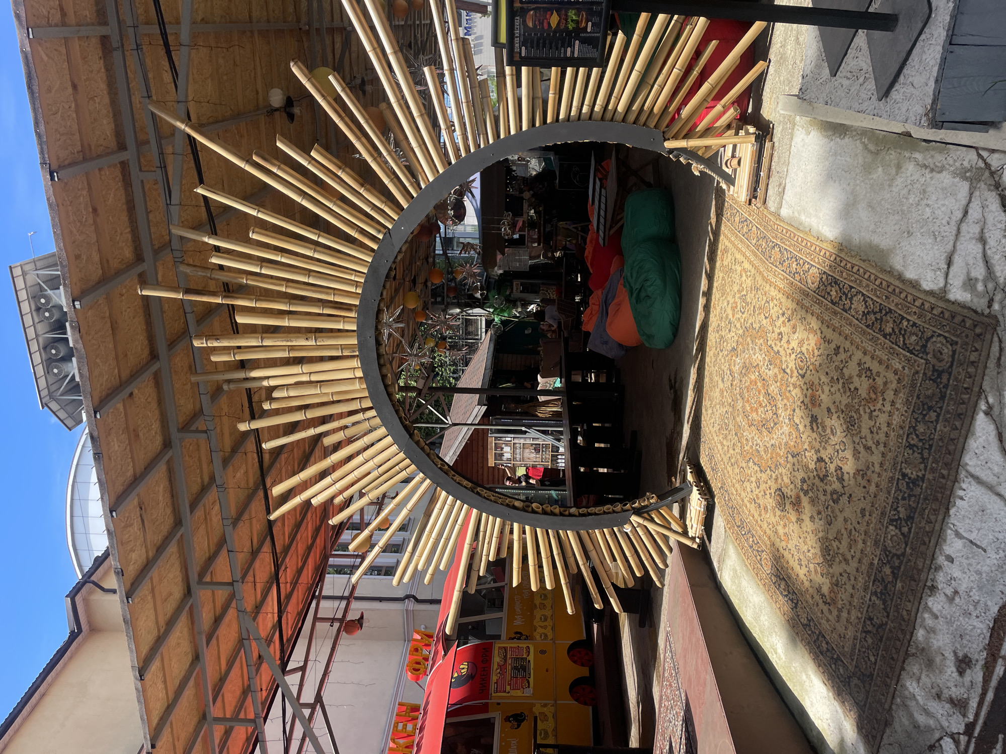 A commercial scene, situated in the city of Sochi, Russia, presents an outdoor establishment with a distinctive bamboo-framed entrance. The entrance is a partial circle, adorned with bamboo stalks that radiate outwards from a dark metal frame. Inside, there are beanbag chairs and other seating arrangements. A patterned rug covers the floor at the entrance. Adjacent to the bamboo structure, a food truck is visible. Above, a covered ceiling structure can be seen. The lighting suggests it is daytime.