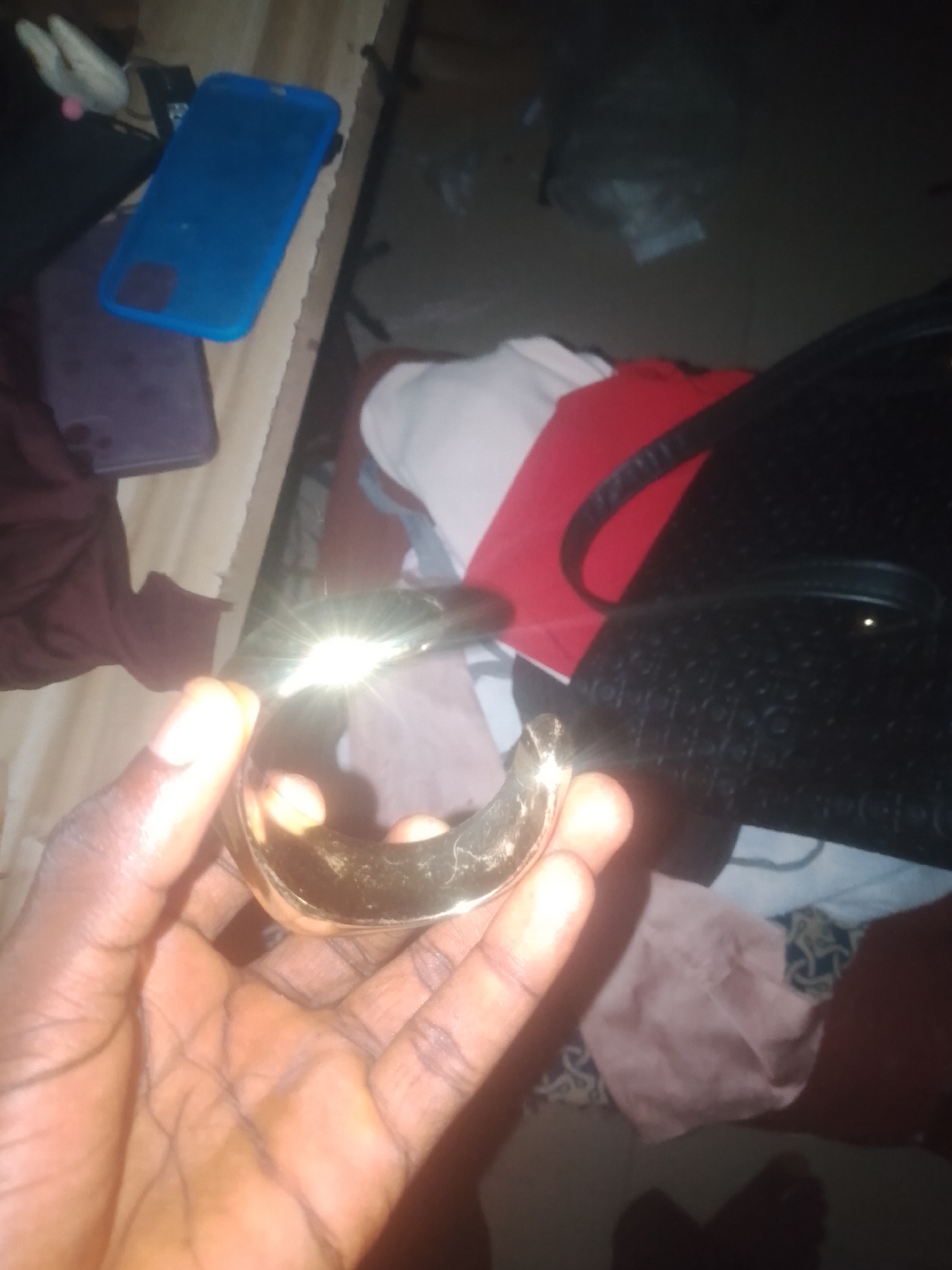 A close-up shot focuses on a person's hand holding a shiny, gold-colored bracelet. The bracelet is curved and appears to be made of metal, with a prominent glint of light reflecting off its surface. The hand holding the bracelet is dark-skinned, with visible fingerprints and skin texture. In the background, various objects are scattered: a blue phone case, a purple phone case, a black handbag with a textured surface, and some articles of clothing in red, white, and dark colors. The environment appears to be indoors, with a wooden surface visible to the left. The lighting is dim, contributing to the overall dark tone of the image. No specific details can be inferred about the location being Jauro Yinu, Nigeria.