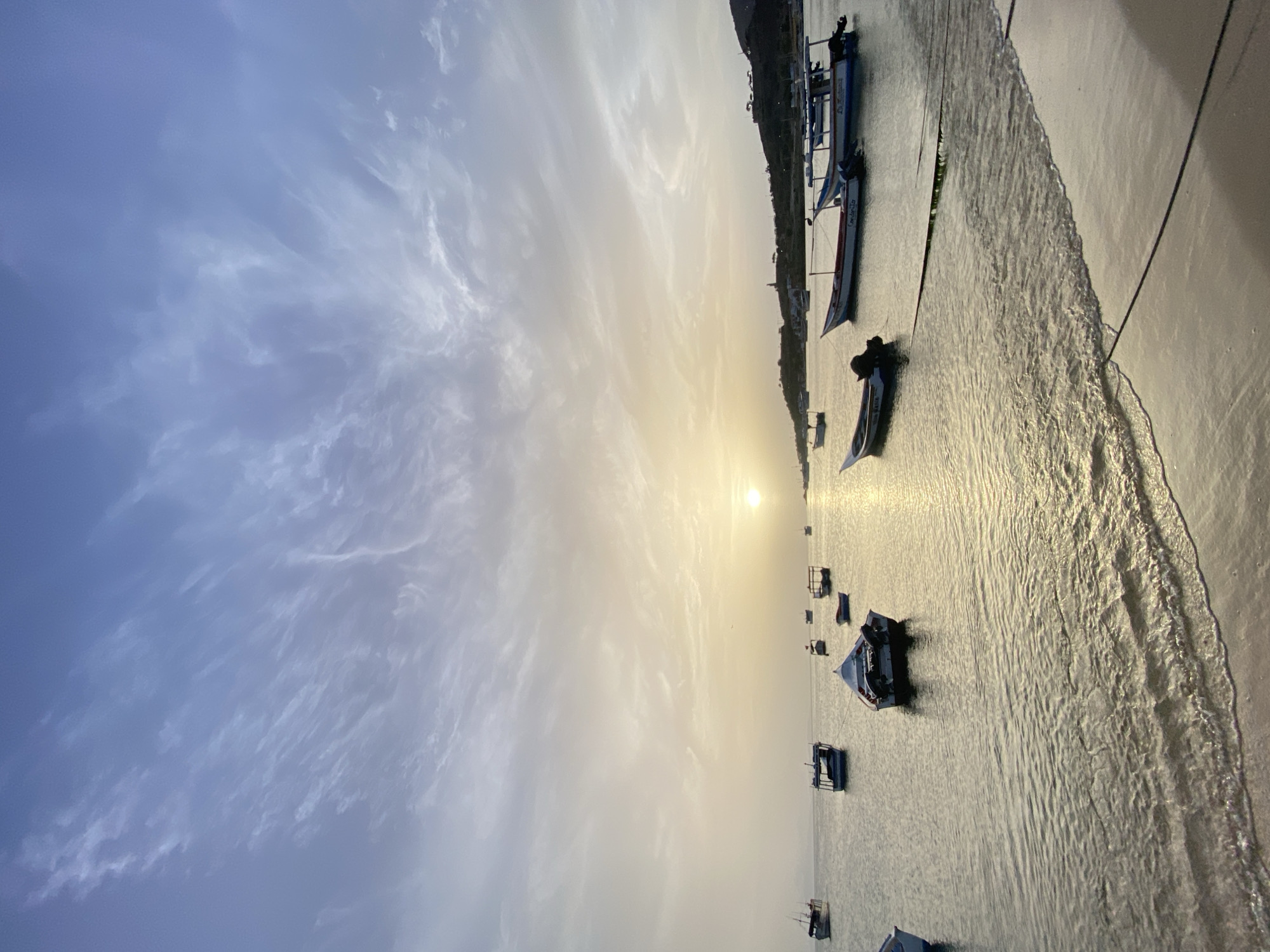 The image depicts a serene coastal scene at Juan Griego, Venezuela, likely during sunset or sunrise. The sky is filled with wispy, streaky clouds in varying shades of blue and white, with a soft, diffused light emanating from the sun, which is low on the horizon. The sun's rays cast a golden glow across the tranquil water, creating shimmering reflections.
Scattered across the calm sea are numerous small fishing boats, some appearing to be at anchor, while others are moored closer to the shore. The boats are varied in design, with some featuring outboard motors and others appearing to be more traditional open vessels. The water laps gently against a sandy beach in the foreground, with ropes from the moored boats extending to the shore. In the background, a hilly coastline is visible, dotted with some structures and vegetation. The overall impression is one of peacefulness and quietude, characteristic of a coastal town at the beginning or end of the day. There are no people visible in the image.