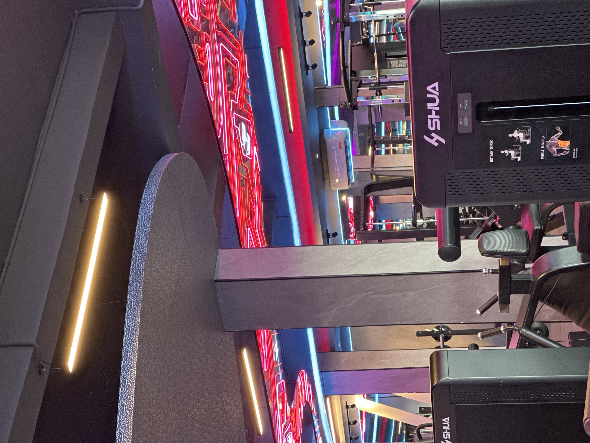 The image depicts a view inside a gym, likely in a modern or upscale setting, potentially in North Kuta, Indonesia. Predominantly visible is a black "SHUA" branded rotary torso exercise machine with instructional diagrams for the user. The gym's interior design is characterized by geometric shapes and vibrant neon lighting in red, blue, and purple hues, arranged in linear patterns that draw the eye into the background. There are support beams of gray, that adds to the architectural aspect.