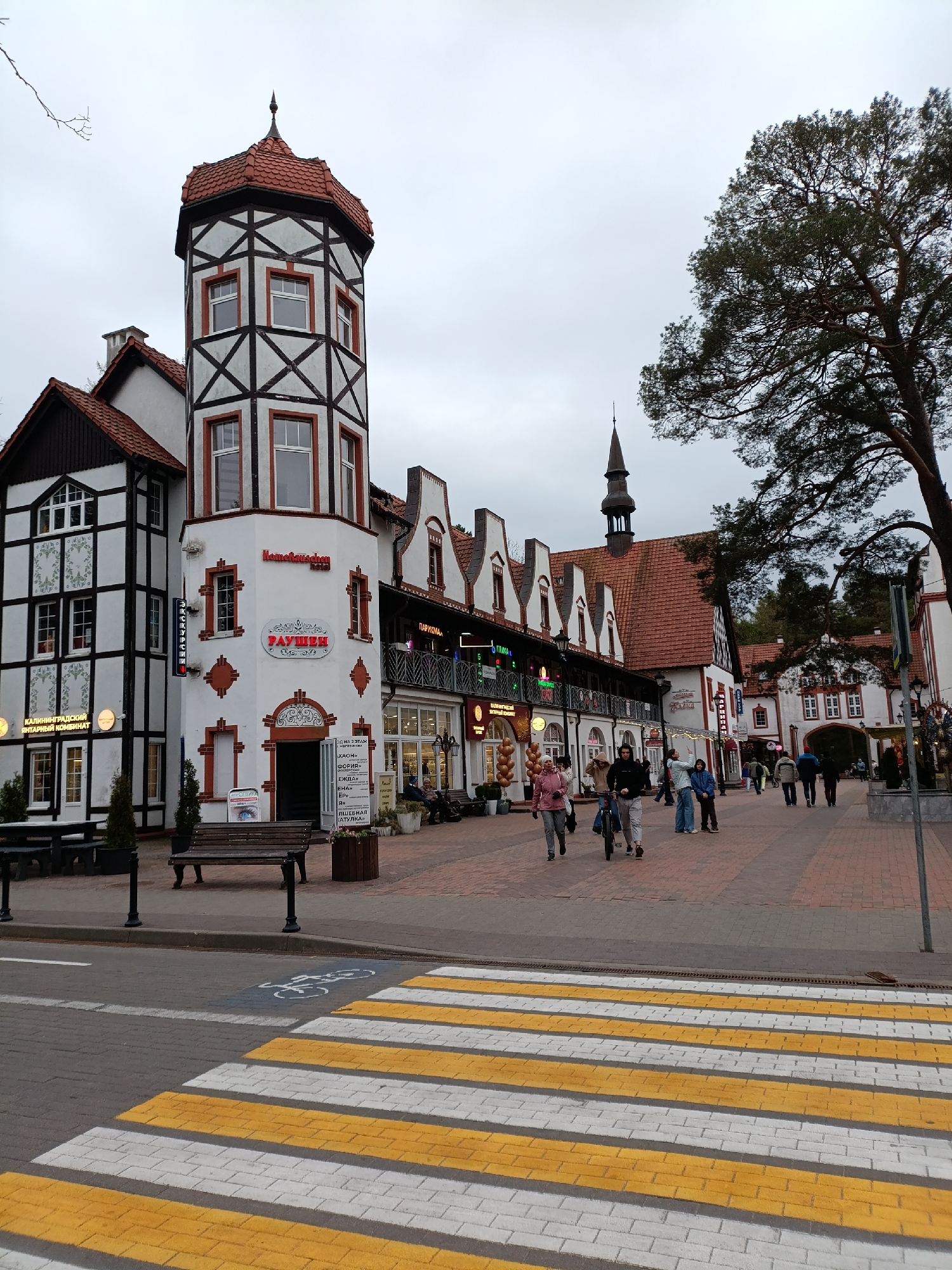 The image captures a street scene in Svetlogorsk, Russia, characterized by distinctive European-style architecture. The building on the left features a tower with half-timbered construction and red-tiled roofing. A storefront displays signage in Russian, including "КАЛИНИНГРАДСКИЙ ЯНТАРНЫЙ КОМБИНАТ" and "РАУШЕН."
A pedestrian crosswalk with yellow and white stripes marks the foreground, while a bench sits near the curb. Several people are walking along the brick-paved pedestrian area. A large tree stands to the right, partially obscuring a church steeple in the distance. The sky is overcast, creating a muted lighting environment.