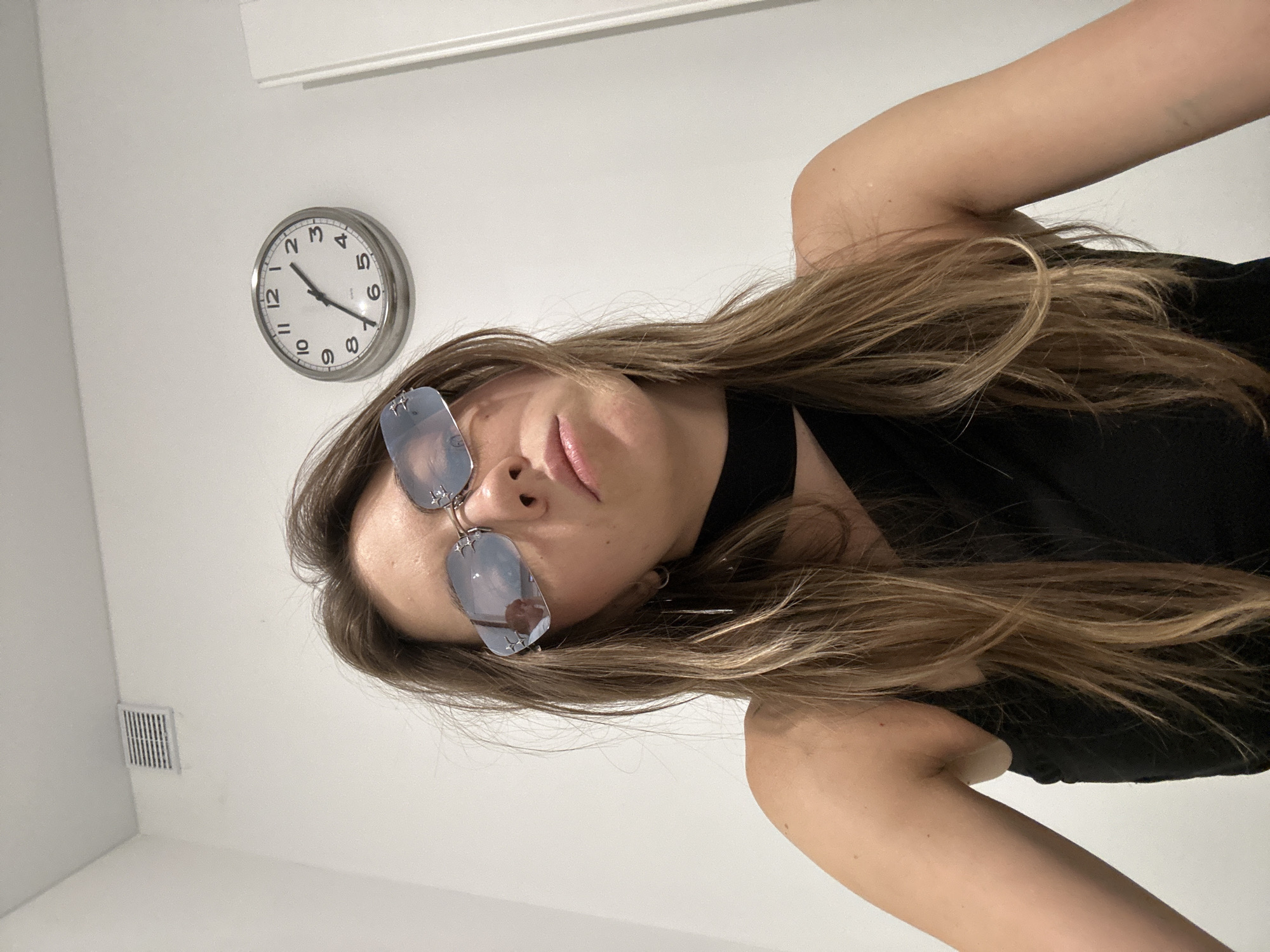A young woman with long, light brown hair is posing for a selfie in an indoor environment. She is wearing sunglasses with a distinctive star-shaped embellishment on the frame, a black choker, and a black top. Her hair is parted in the middle and falls over her shoulders. The background consists of a white wall with a ventilation grate and a round clock on the wall. There is also a sliver of a white door on the right side of the frame. The lighting appears to be artificial. The image was captured at an upward angle. There is no identifiable information or context suggesting the location is Moscow, Russia.