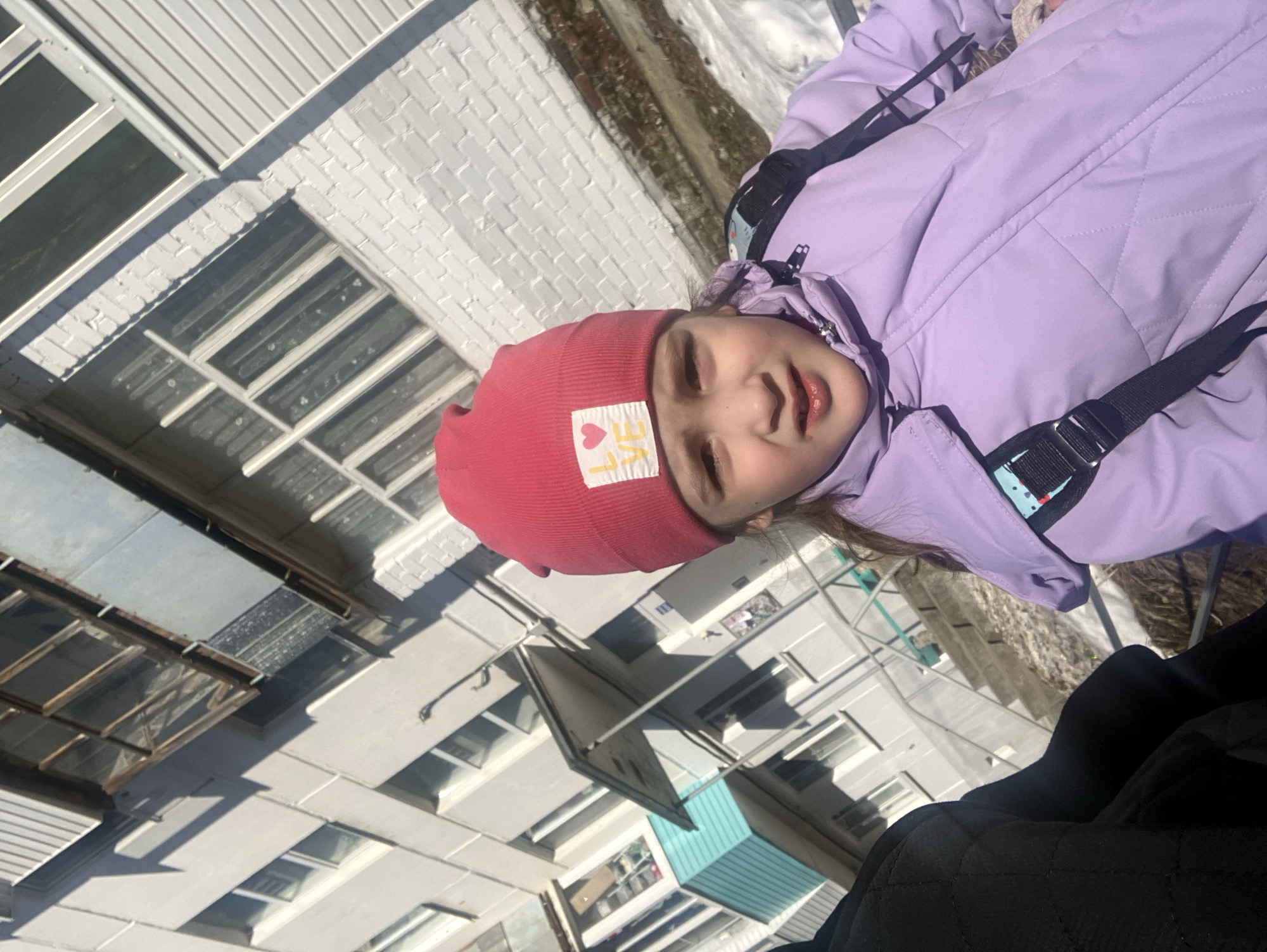 The image shows a child wearing a pink hat and a lavender jacket. The background consists of buildings, which are consistent with architecture found in Chaikovsky, Russia. Snow is present.