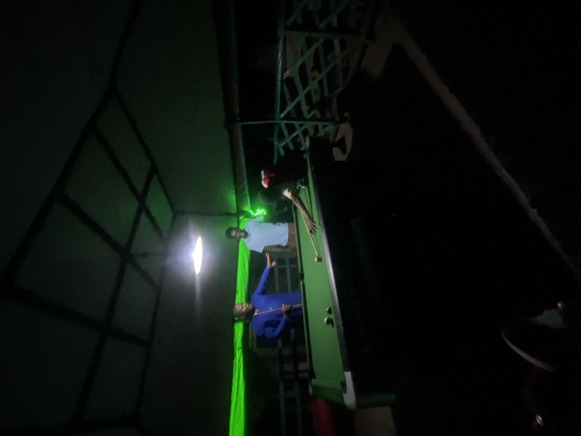 The scene is located in Lokoja, Nigeria, within a dimly lit, covered recreational space. Three adult males are present around a green-felted pool table. A single bright, unshielded light source hangs centrally above the pool table, illuminating the playing surface, while green LED strip lighting casts a distinct green glow on the upper walls and ceiling perimeter.
A male subject, wearing a blue long-sleeved collared shirt, stands to the left of the pool table. He holds a pool cue in his right hand and extends his left arm with an open hand, pointing towards the table or another individual. To his right and slightly behind him, a second male subject, dressed in a light blue short-sleeved collared shirt and light brown trousers, stands observing the table. A third male subject, wearing a light-colored top and a red head covering, leans over the right side of the pool table, preparing to strike a white cue ball with a pool cue. At least two other colored pool balls (yellow and green/blue) are visible on the table surface.
The ceiling features a dark, grid-like structural pattern, and a dark green railing or lattice structure is visible in the background on the right side of the image. The immediate foreground and lower portions of the room are predominantly dark, with indistinct flooring. The individuals are engaged in or observing a game of billiards.