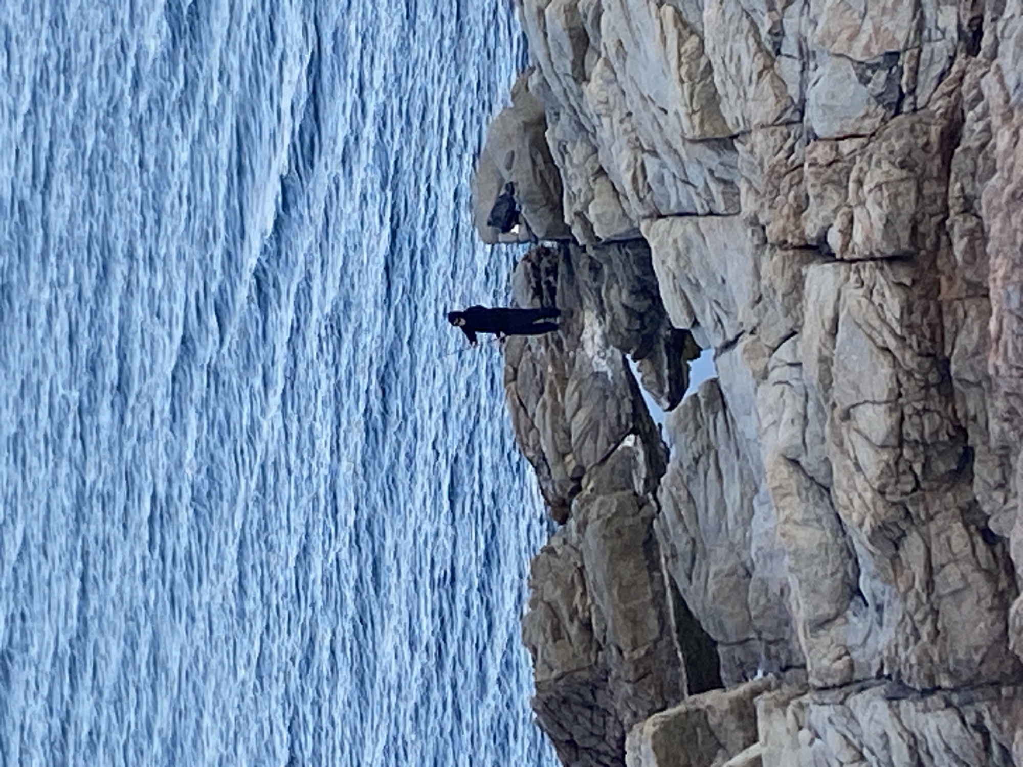 The image shows a person tightrope walking on a rope stretched between rocky cliffs near the sea. The person is dressed in black and appears to be using a balancing pole. The sea is visible on the left. The location is most likely in Palafrugell, Spain, given the prompt's instructions.
