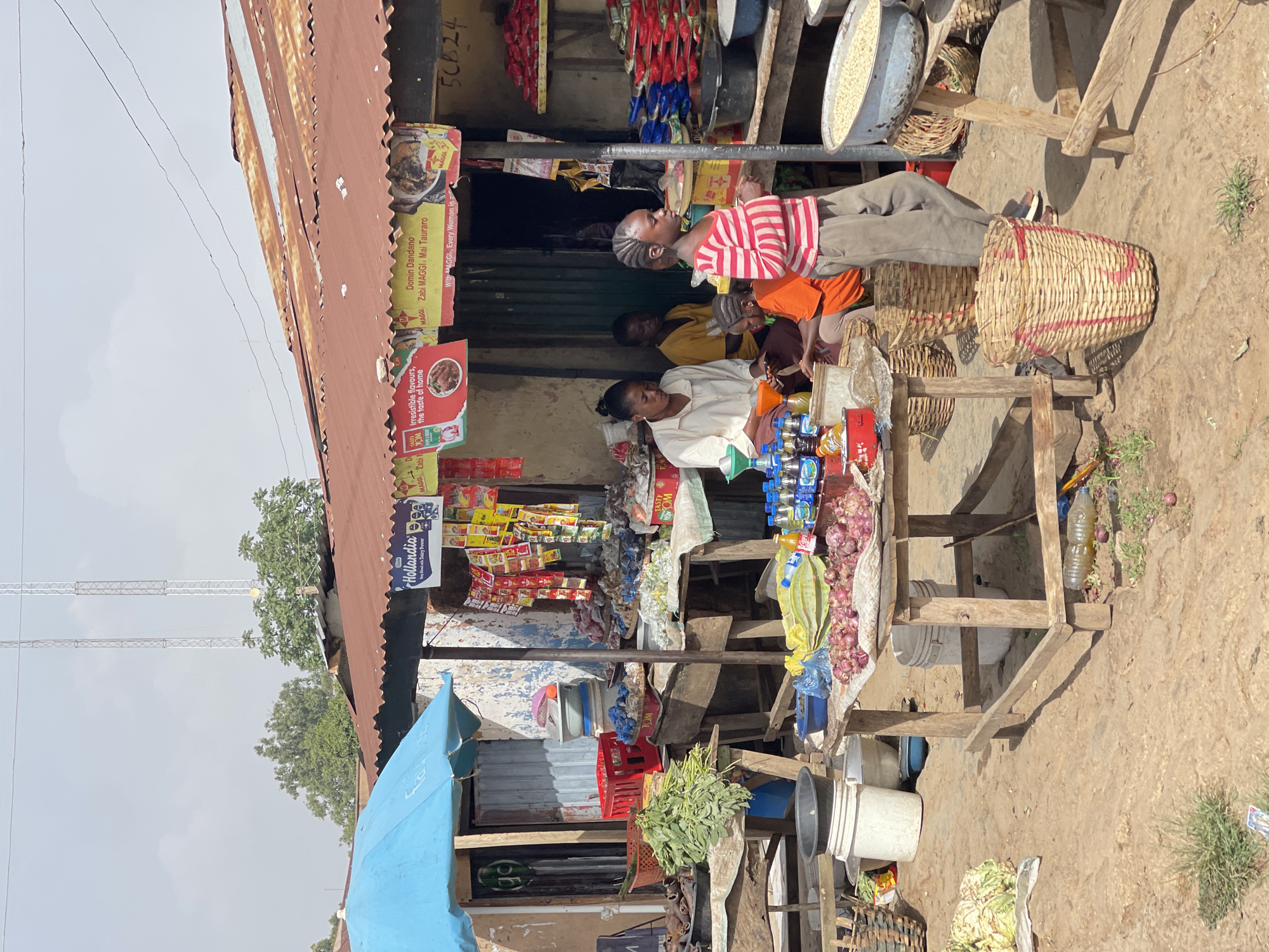The image captures a bustling scene at a roadside market stall in Jos, Nigeria. The stall is constructed from weathered wood and corrugated metal, displaying a variety of goods for sale. There are several people in the image. An adult woman sits behind the stall, attending to the items for sale. A younger individual wearing a striped shirt is standing next to the stall. There are additional people seated within the shaded area of the stall.
The stall offers a diverse range of products, including packets of spices and seasonings, drinks in bottles, onions, and fresh green vegetables. Several buckets and baskets, some filled with produce, are positioned around the base of the stall.
Posters and signs advertising products, such as Hollandia dairy and Maggi seasoning, are affixed to the structure of the stall. Towering communication towers are visible in the background, indicating the presence of urban infrastructure. The ground surrounding the stall is dusty and uneven.