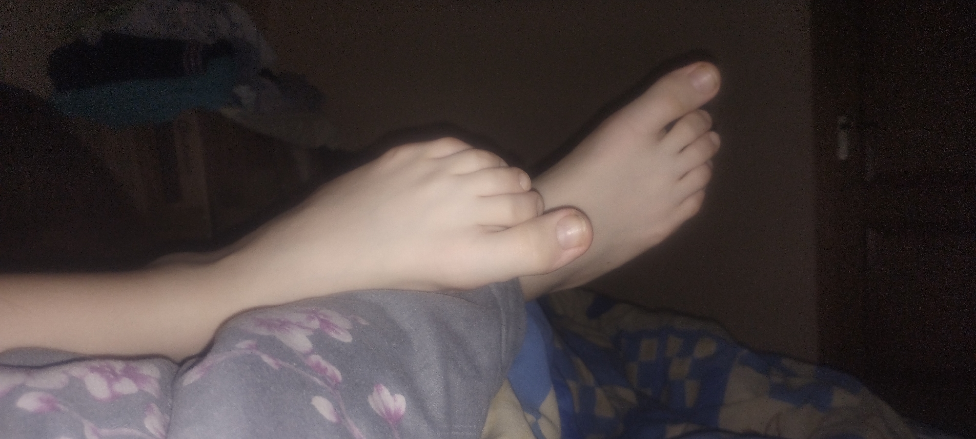 The image shows a close-up view of two human feet, likely belonging to a child given their size and proportions. The feet are resting on a gray blanket with a floral pattern in shades of purple and pink. The background is dark, suggesting an indoor setting, possibly a bedroom. One foot is positioned in front of the other, with the toes of the front foot clearly visible. The toenails appear to be unpolished. The lighting is dim, casting soft shadows and highlighting the pale skin of the feet. There is no discernible information within the image to indicate a specific location in Zaporizhzhia, Ukraine.