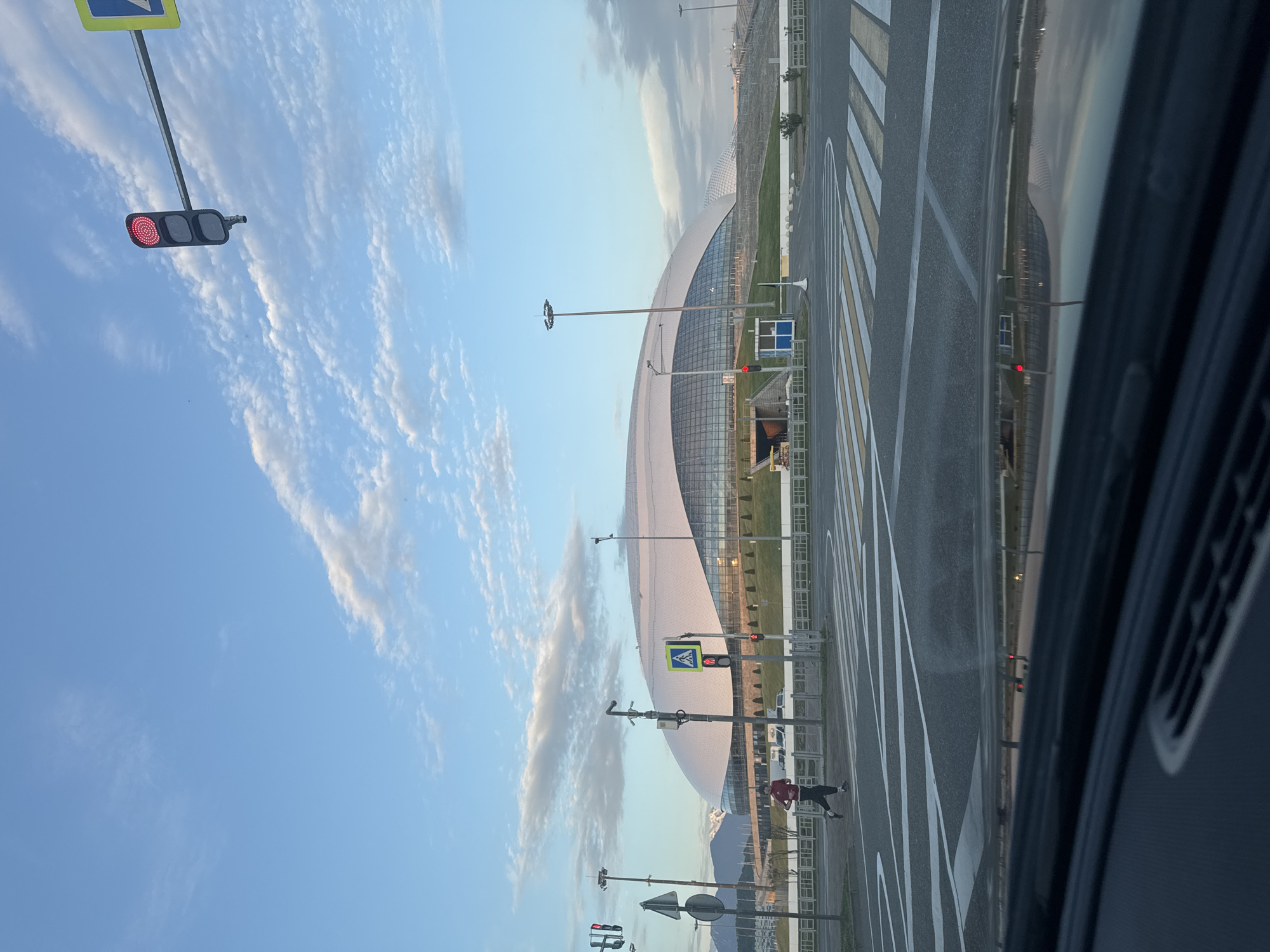 The image depicts a view from inside a vehicle, looking out towards a stadium in the city of Sirius, Russia. The sky is partly cloudy, with patches of blue visible. A traffic light shows red. A pedestrian is visible on the sidewalk near the crosswalk. Striped markings indicate crosswalks across the road. The stadium has a distinctive curved, translucent roof structure. Streetlights line the road. A mountain range is visible in the background.