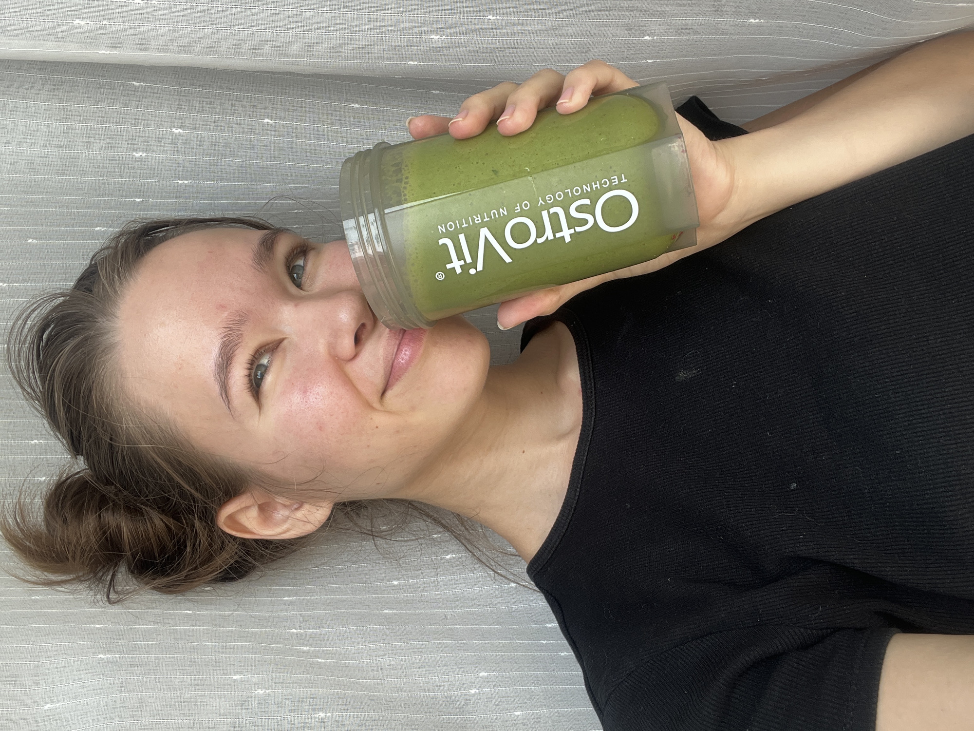A young woman lies on a light gray horizontally-striped surface. She holds a plastic container of green smoothie in her right hand near her face. The container has the word "OstroVit" and "TECHNOLOGY OF NUTRITION" printed on it in white letters. She has light skin, brown hair pulled back into a loose bun, and is wearing a black shirt. The environment appears to be indoors. The location is not discernible from the image.
