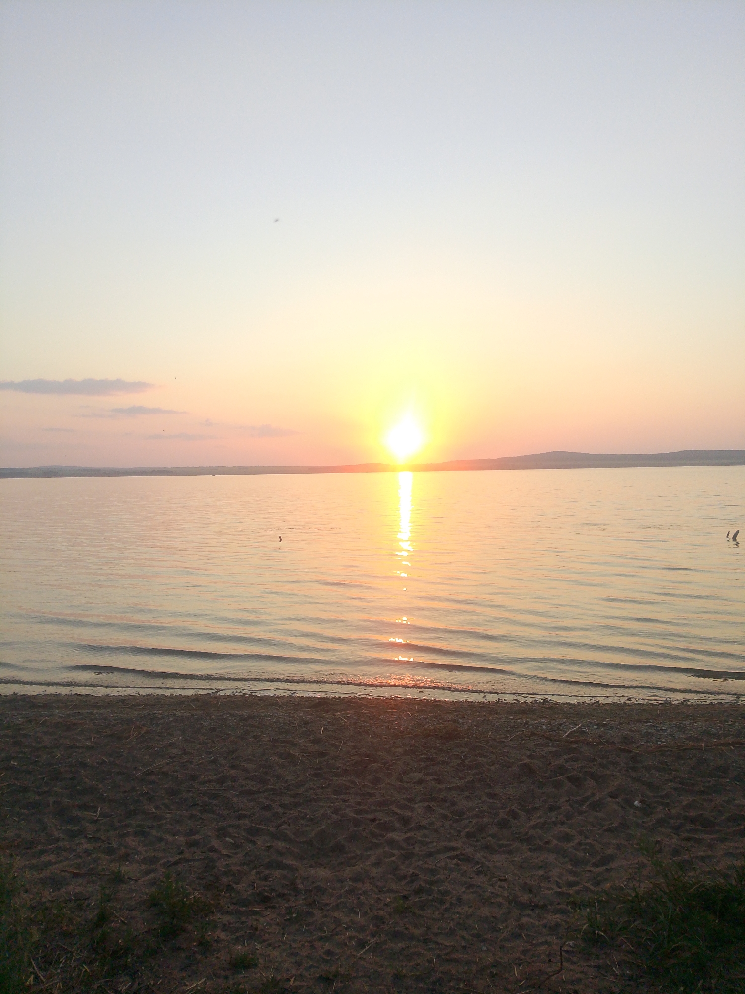 The image depicts a serene sunset over a body of water, likely a lake or sea, with a sandy beach in the foreground. The sun, a bright orb, is positioned near the horizon, casting a warm, golden glow across the water and the sky. Gentle waves lap at the shore, creating a rhythmic pattern on the water's surface. The sky transitions from a pale blue at the top to hues of orange and yellow near the sun, with a few scattered clouds. The beach appears to be composed of sand and small pebbles, with some sparse vegetation growing along the edge. There are no visible people or specific landmarks that would definitively place this scene in Mnogosopochnoye, Kazakhstan. The overall atmosphere is peaceful and natural.