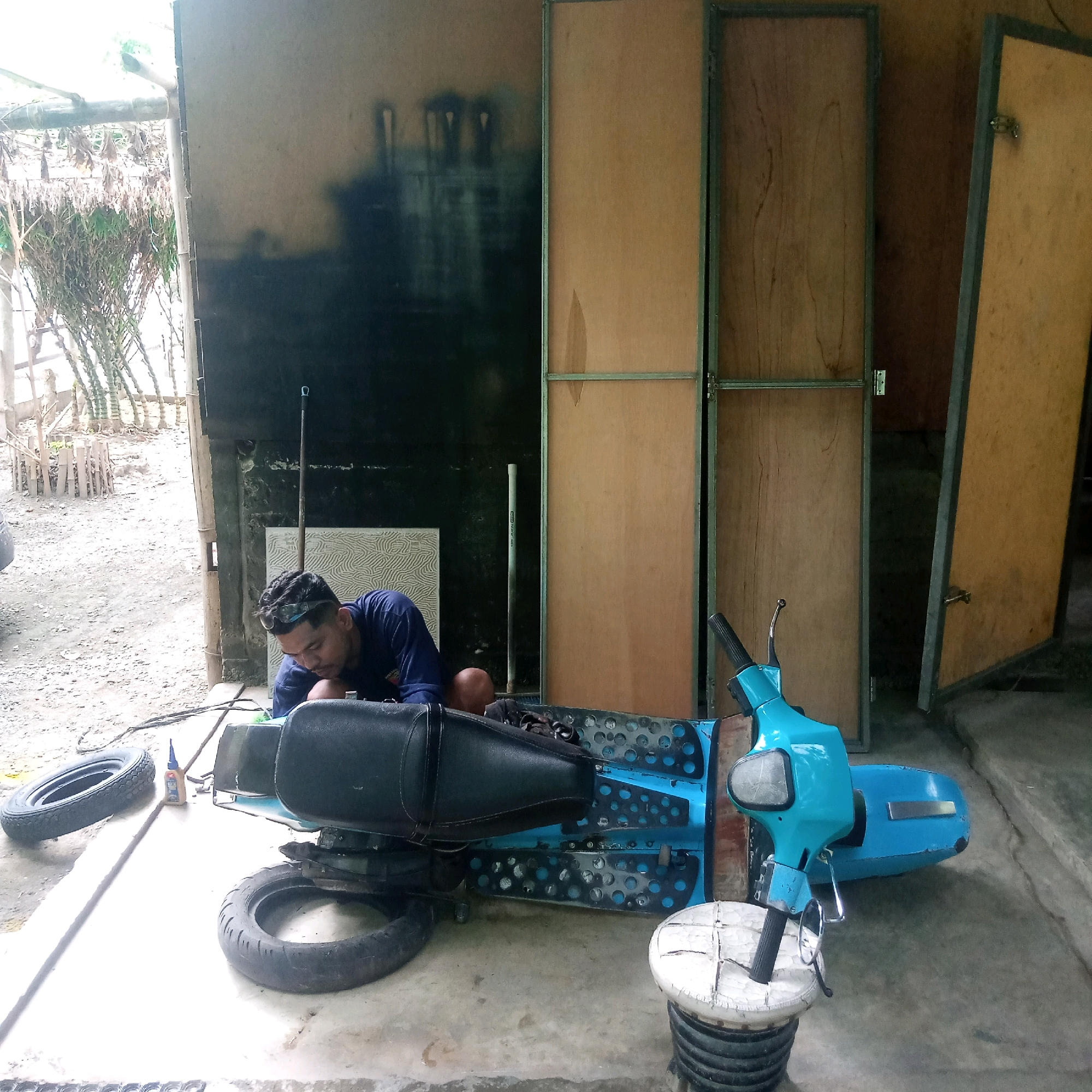 An adult male with dark hair and a blue shirt is working on a turquoise scooter in an outdoor, open-air workshop. He is wearing safety glasses pushed up on his forehead. The scooter's body is disassembled, with the seat off and the chassis exposed. A tire lies to the left of the scooter and another is positioned under the chassis. A small bottle of glue is also on the ground near the tire. The workshop appears to be constructed with a wooden frame and some corrugated metal sheeting. The background shows a dirt ground area with some bamboo plants and wooden fencing. The walls behind the scooter are made of plywood panels. The scene is lit by natural daylight.
