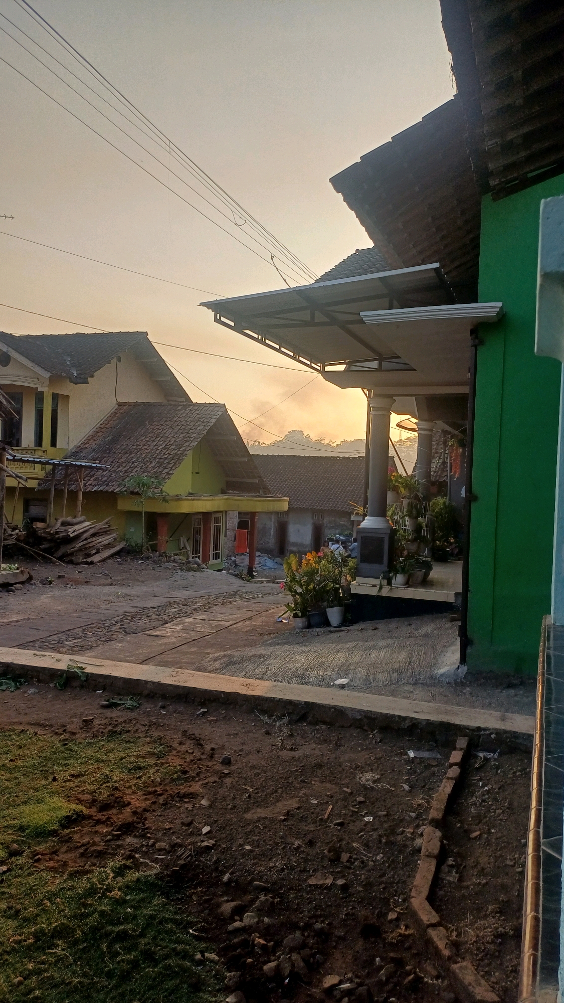 The image depicts a residential scene in Wonokerso, Indonesia, during what appears to be early morning or late afternoon, given the soft, warm lighting and hazy sky. A multi-story house with a yellow facade and a tiled roof is visible on the left, with a smaller, yellow single-story structure adjacent to it, also with a tiled roof. Construction materials, including stacks of wood, are piled near the yellow structures.
To the right, a building with a prominent green facade features a covered walkway with a white awning supported by metal beams. This walkway leads to an entrance with stone-clad pillars and multiple potted plants. The ground in the foreground is a mix of patchy grass, dark soil with scattered rocks, and a concrete pathway or patio area. Electrical wires are strung across the sky, and distant hills or mountains are faintly visible through the haze. The overall impression is one of a rural or semi-rural residential setting undergoing some form of construction or renovation.