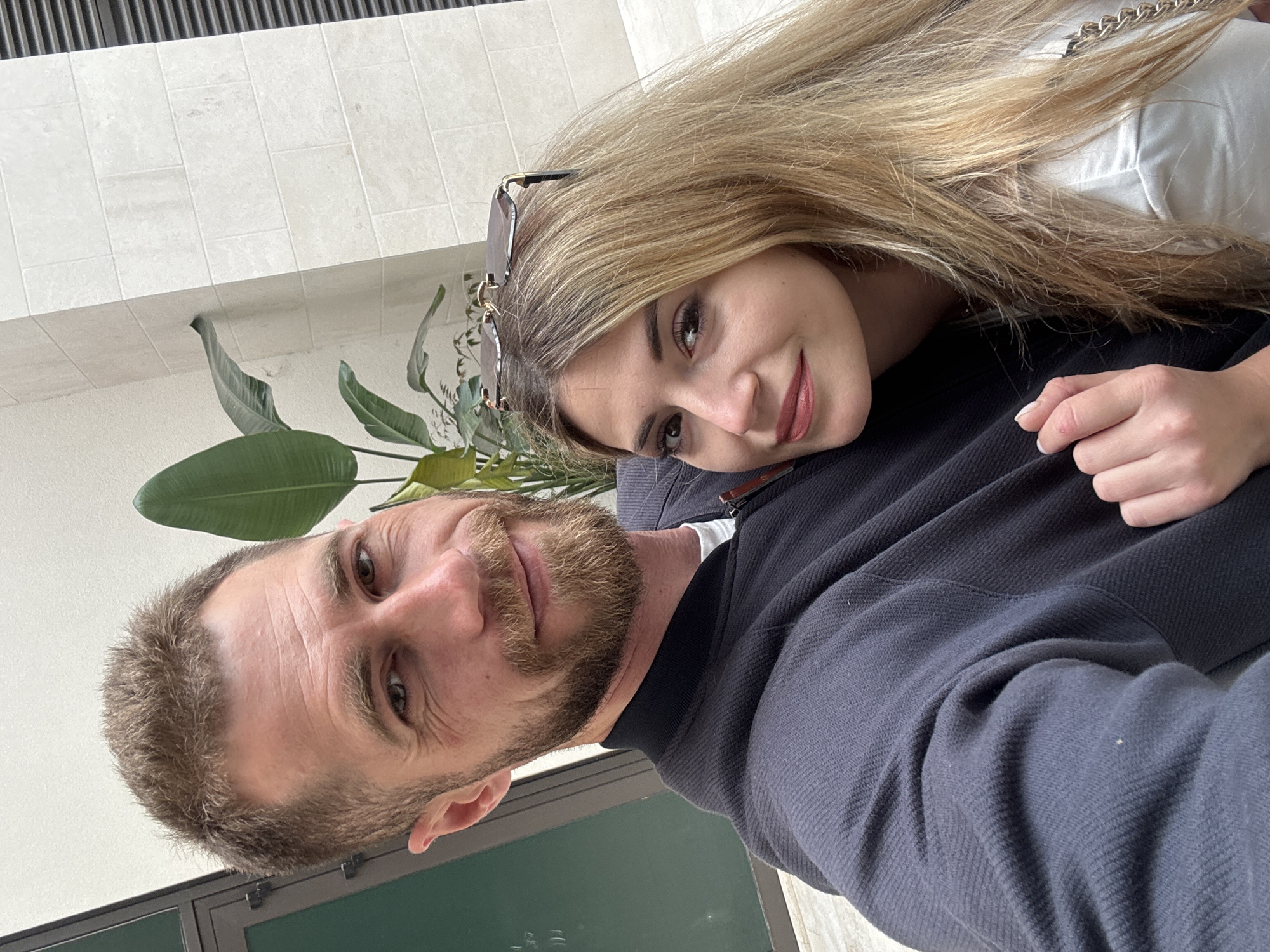 A selfie captured from a low angle features a man and woman embracing near a building with a tiled white facade. The man, positioned in the foreground, has short brown hair and a beard. The woman, lying on his chest, has long blonde hair and sunglasses resting on her head. A potted plant with large green leaves is visible in the background. The window frame, constructed of dark metal, is also visible in the bottom left. The location is likely indoors, near a window or entrance of a building in Tivat, Montenegro.