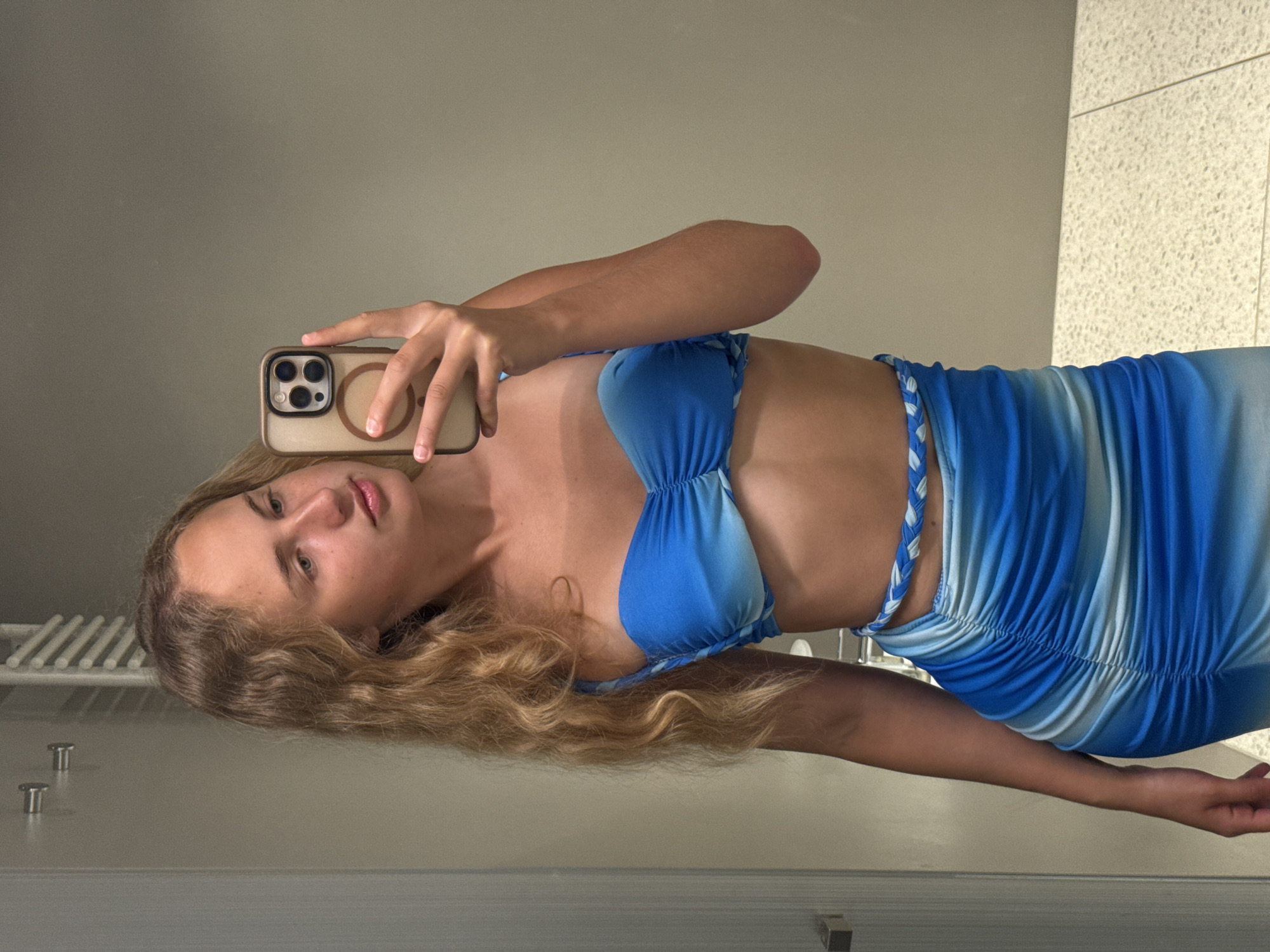 A young woman with blonde, wavy hair takes a selfie in a bathroom. She is wearing a blue ruched bikini top and a matching blue ruched skirt with a braided belt. Her phone is held up to take the photo, and her right hand is visible. The background appears to be a wall with a towel rack, a door handle, and tiled flooring. The lighting suggests an indoor environment, possibly a hotel bathroom.