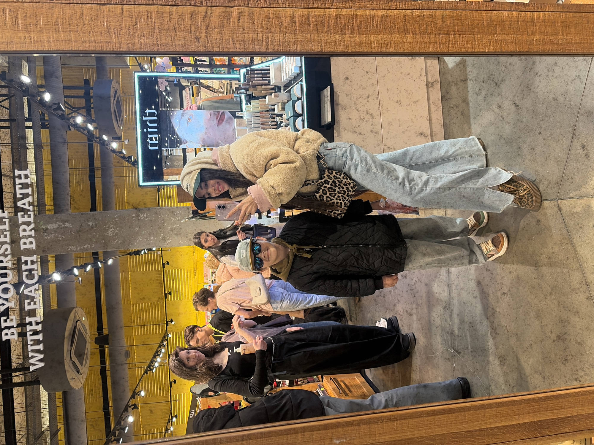 The image shows a reflection in a mirror, seemingly inside a retail space. A group of people is visible, positioned near the mirror. One woman is holding a phone, possibly taking a picture of the group. Another woman is wearing a beige, Sherpa-style jacket and light-colored jeans. Several other individuals are partially visible, their attire varying from black clothing to lighter colors. The store features lighted displays. Outside the retail space, a sign reads "BE YOURSELF WITH EACH BREATH." Based on the store signage, the location is likely related to the "Rehab." brand. The exact city of Saint Petersburg, Russia, cannot be definitively determined from the image alone without additional visual information or metadata.