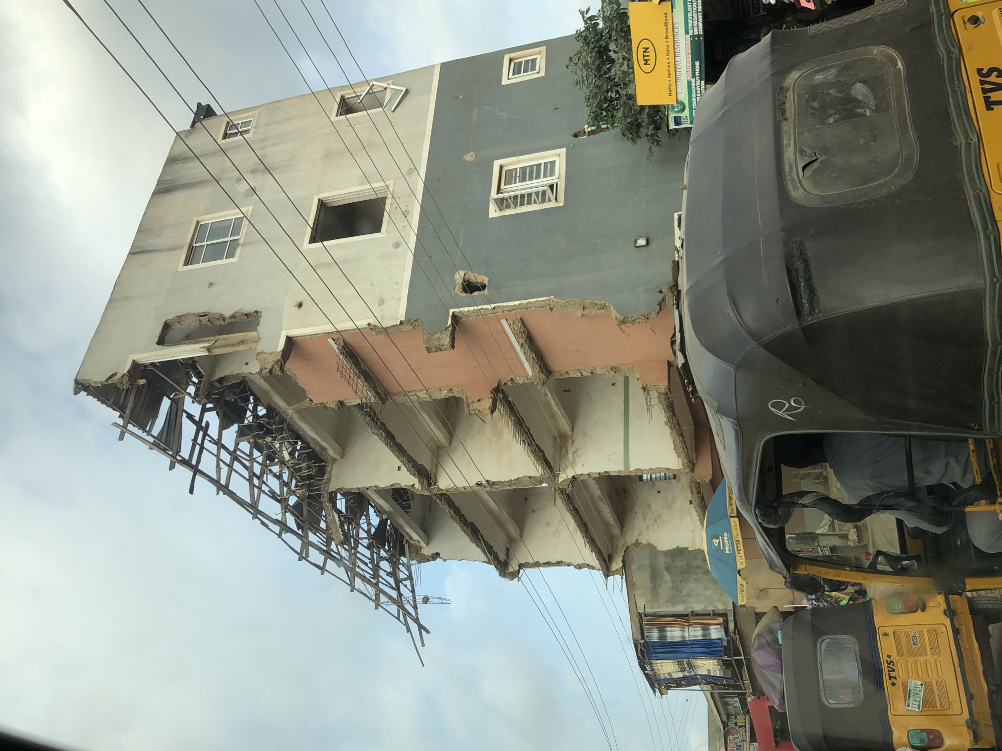 The image depicts a severely damaged multi-story building in Otta, Nigeria, with its roof and several upper floors collapsed. The structural integrity of the building is compromised, exposing internal concrete and rebar. Various building materials, including wood, metal, and debris, are scattered around the structure. Several windows are intact, while others are missing or broken. Power lines crisscross the scene. In the foreground, a yellow auto-rickshaw (tricycle) is visible, and the back of another auto-rickshaw can be seen to its left. The sky is overcast, suggesting a cloudy day. A yellow sign with the MTN logo is mounted on a pole near the damaged building. The overall scene conveys destruction and disarray.