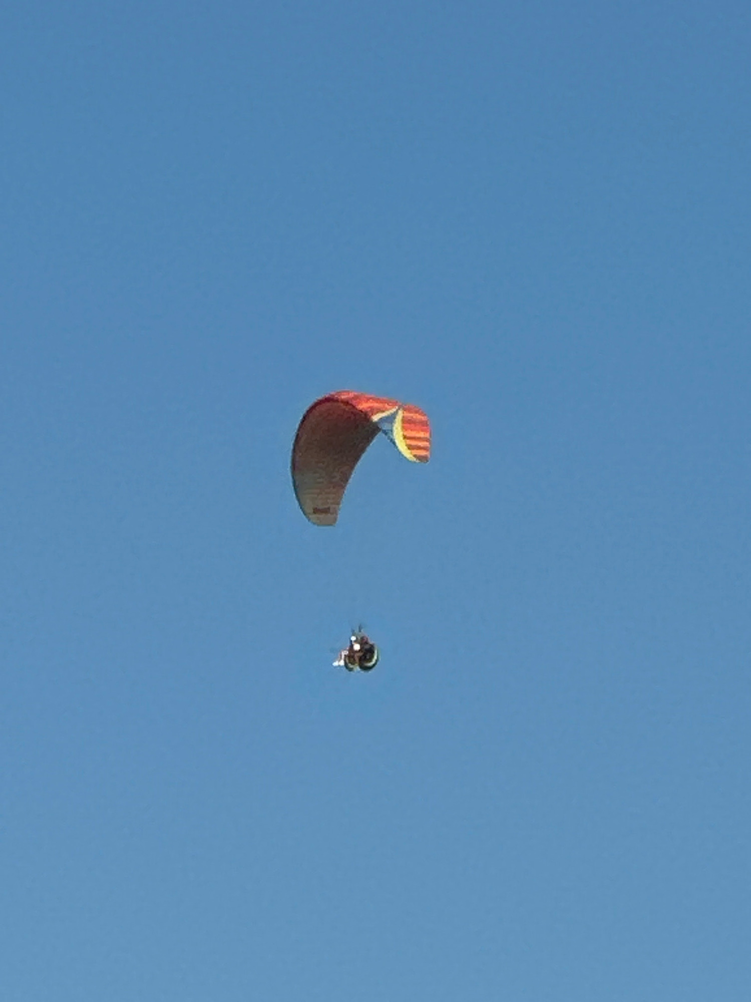 A paraglider is in mid-air against a clear blue sky. The paraglider's wing is colorful, with red, orange, and yellow stripes. A person is attached to the wing, suspended beneath it. The image appears to have been taken during daylight hours. No specific location in the city of Boreti, Montenegro is identifiable from the image.