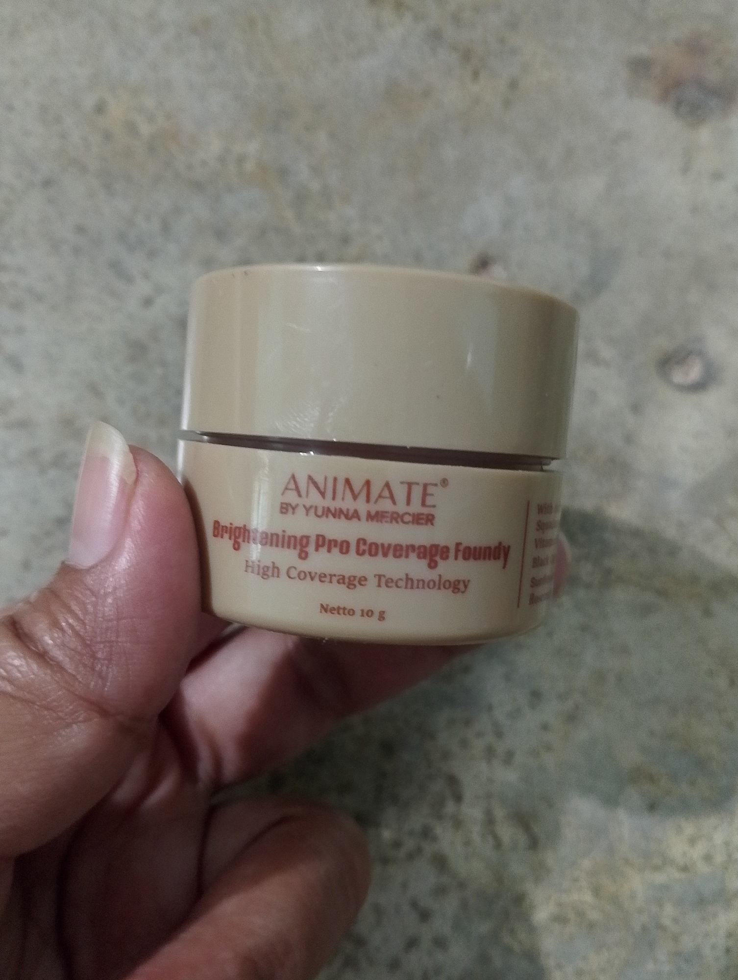 A person's hand is holding a small, light-colored jar of "ANIMATE BY YUNNA MERCIER Brightening Pro Coverage Foundy High Coverage Technology Netto 10 g". The jar is cylindrical with a threaded lid, and the text is printed in red. The background is a textured, light-colored surface, possibly a wall or floor. The lighting is natural, and there are no prominent shadows. The image appears to have been taken indoors, or in a shaded outdoor area, and the focus is on the product.
