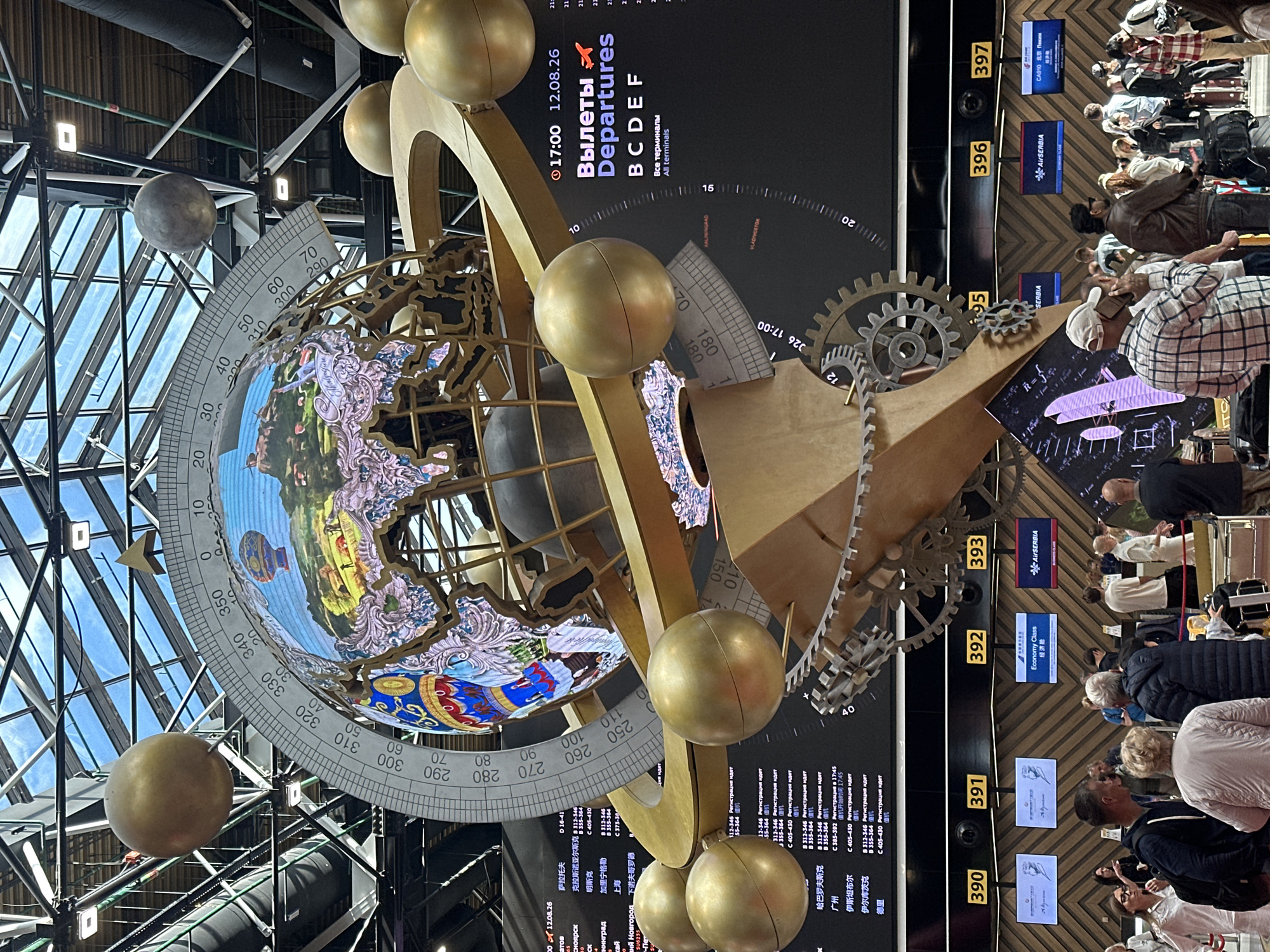 The image depicts the interior of a modern airport terminal in Khimki, Russia, likely Sheremetyevo International Airport, identifiable by its architectural style and departure board information. In the foreground, a large, elaborate sculpture resembling a globe with orbiting golden spheres and metallic gears dominates the space. The globe itself is a detailed LED display showing a vibrant landscape with hot air balloons and buildings. Behind the sculpture, a large digital departure board lists flight information, with Russian text indicating destinations and flight numbers. The board displays "Вылеты Departures" and lists cities such as "Москва," "Пунта-Кана," "Нью-Йорк," "Шанхай," and "Хабаровск." Numerous people, dressed casually, are seen standing and walking in the terminal, many with luggage, suggesting they are travelers. Some individuals are looking at their phones, talking, or queuing. The environment is well-lit, with a high ceiling featuring a transparent roof that allows natural light to filter in, highlighting the overall spacious and busy atmosphere of an international airport. Signs for "Economy Class" and airlines like "AIRSERBIA" are visible, further confirming the airport setting. The overall scene captures the dynamic environment of air travel, emphasizing both the infrastructure and the human element.
