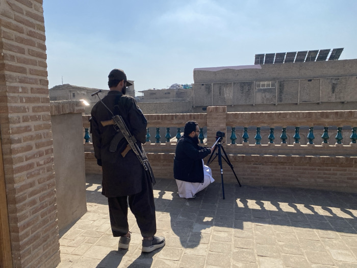 This image depicts an elevated outdoor terrace or rooftop in Kandahar, Afghanistan, under a clear daylight sky.
In the foreground-left, a male figure stands with his back partially to the viewer, wearing dark long-sleeved clothing, dark trousers, dark shoes, and a dark baseball cap. He has a dark beard. An AK-style assault rifle, featuring a wooden stock, is slung over his right shoulder.
To the right of this individual, a second male figure is crouched, operating a black camera mounted on a black tripod. This individual wears a dark jacket over white traditional attire, glasses, and has a dark beard. The camera is oriented towards the background.
A reddish-brown brick balustrade with dark green vertical ornamental elements separates the immediate foreground from the middle ground. The floor of the terrace is paved with light-colored rectangular tiles, showing distinct shadows cast by the balustrade and the standing figure, indicative of direct sunlight.
In the background, under a clear blue sky, numerous urban buildings are visible. One prominent building features a series of multiple solar panels mounted on its rooftop.