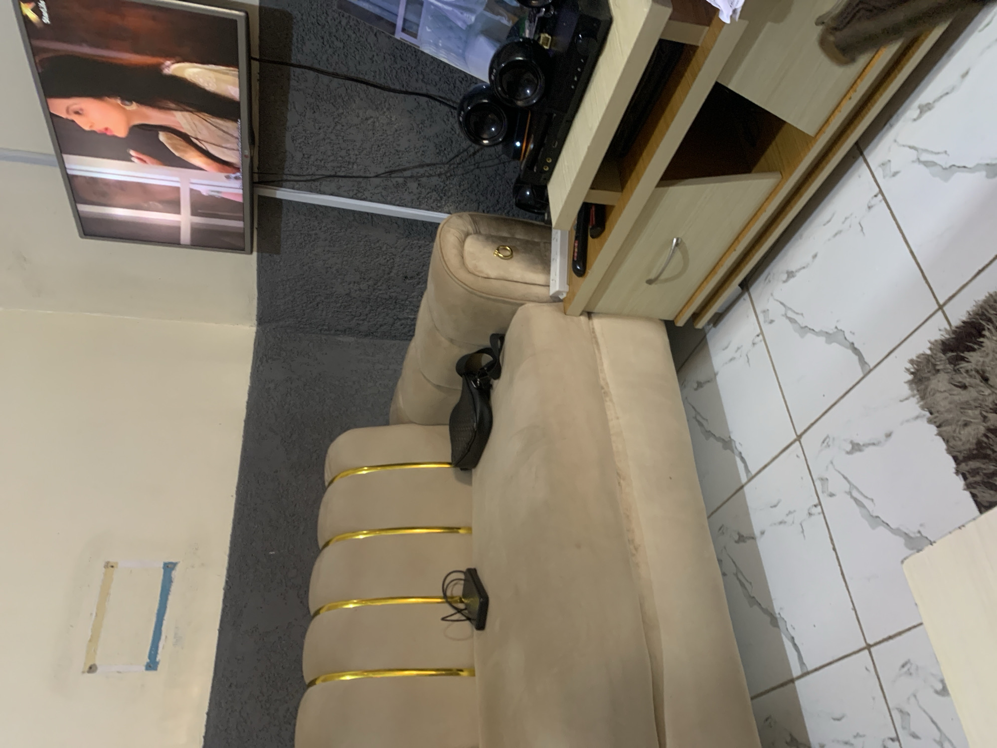 The image depicts an indoor living room scene, possibly located in Jos, Nigeria. In the background, a television is mounted on a stand, displaying a woman. To the left, a beige sofa with gold accents is partially visible. On the sofa, a black object, possibly a charger or power bank, and a black purse are placed. In the foreground, a light-colored entertainment unit with drawers and shelves is situated, holding a black DVD player or sound system with speakers. The floor is tiled with a marble-like pattern. A gray textured wall runs along the top half of the room, contrasting with a lighter-colored wall above it. A partially painted rectangular outline is visible on the upper wall. The overall impression is of a functional, modest living space.