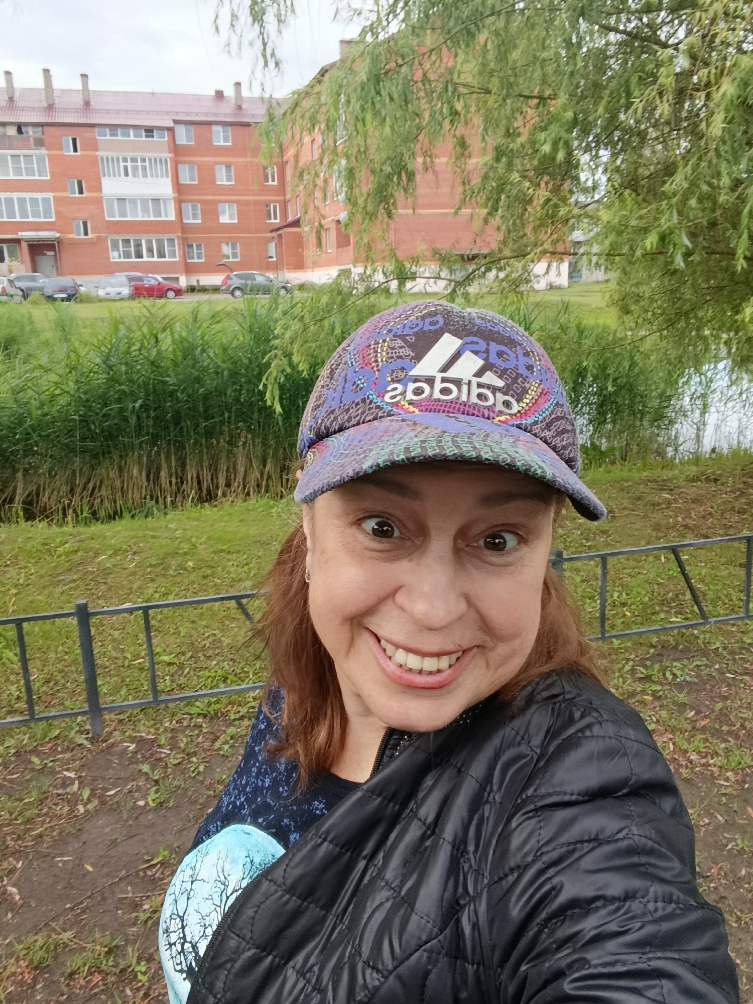 A woman with brown hair and light skin is taking a selfie outdoors. She is wearing a black quilted jacket and a colorful baseball cap with an Adidas logo. She is smiling broadly, showing her teeth. In the background, there is a brick apartment building with several windows and a balcony. Adjacent to the building are green reeds and trees, and a body of water. A metal fence runs across the lower part of the image, separating the foreground from the background. The scene appears to be in a residential area.