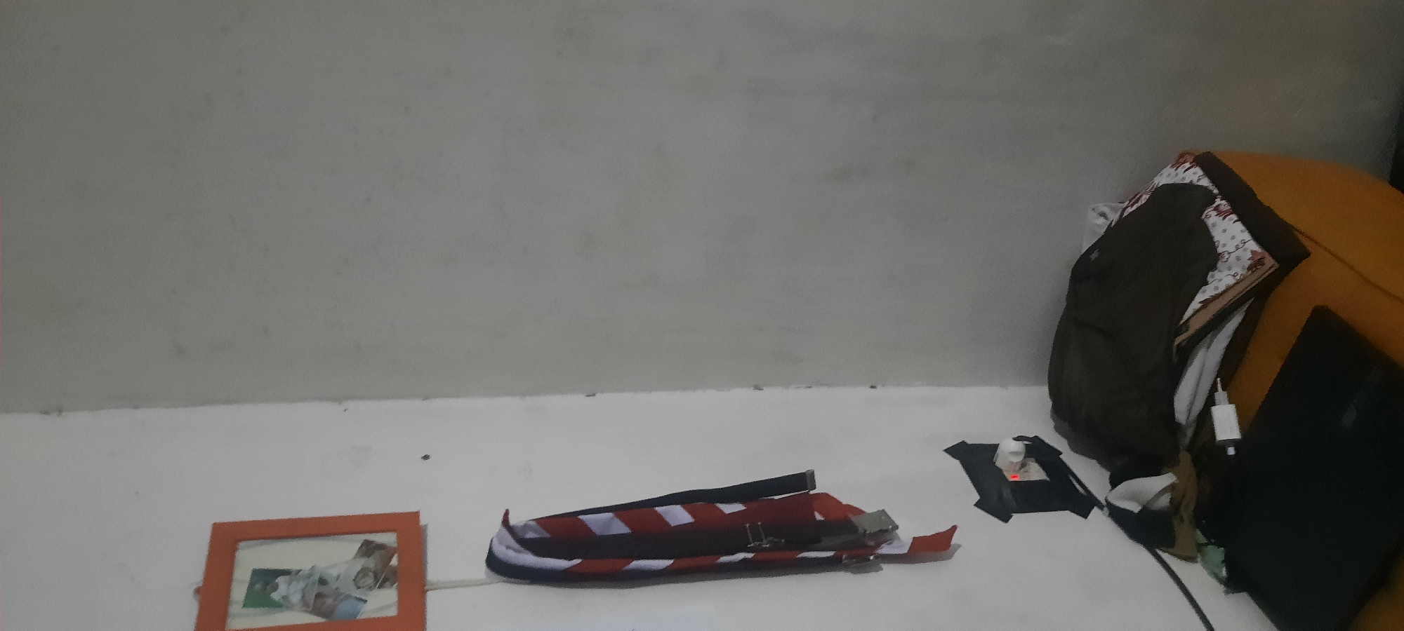 The image depicts a corner of a room with white walls. On the left wall, an orange-framed picture hangs at the top. Below the picture, two belts are suspended. One belt has a striped pattern of red and white, with black accents, and a silver buckle. The other belt is black with a silver buckle. Adjacent to the belts, a white electrical outlet is visible, partially covered by black tape. A small red light is illuminated on the outlet.
On the right side of the image, a yellow upholstered furniture item, possibly a chair or ottoman, is present. A pile of dark clothing, possibly a jacket and shirts, is stacked on the furniture. To the left of the stacked clothing, a black laptop is placed on a flat surface. The environment appears to be an indoor setting within a residential or dormitory-like space. There are no people visible in the image. The lighting is dim, suggesting an interior space with limited natural light. The image quality is grainy, indicating it was likely taken with a low-resolution camera or under poor lighting conditions.
The context of North Jakarta, Indonesia, is not directly discernible from the visual elements of the image itself. The scene shows common household objects and a basic room setup, which could be found in many locations.