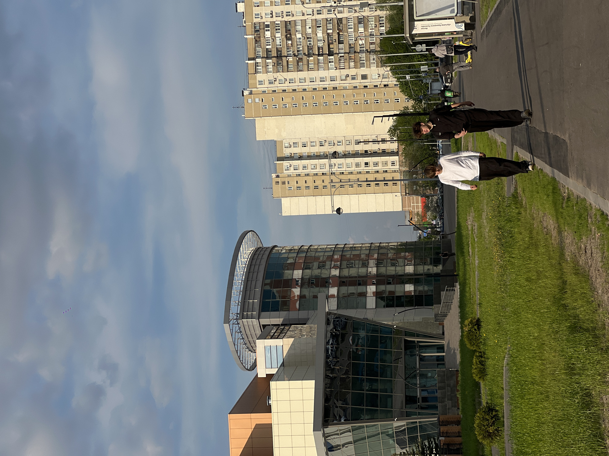 The image shows a cityscape in Moscow, Russia. Two young men walk on a paved walkway alongside a grassy area. One is wearing a white shirt and dark pants, while the other is wearing a black jumpsuit. In the background, there are two distinct buildings: a modern building with a rounded top and reflective windows, and a tall, Soviet-era apartment building. Overhead, there is a blue sky with some clouds and a group of red and purple balloons drifting in the distance.