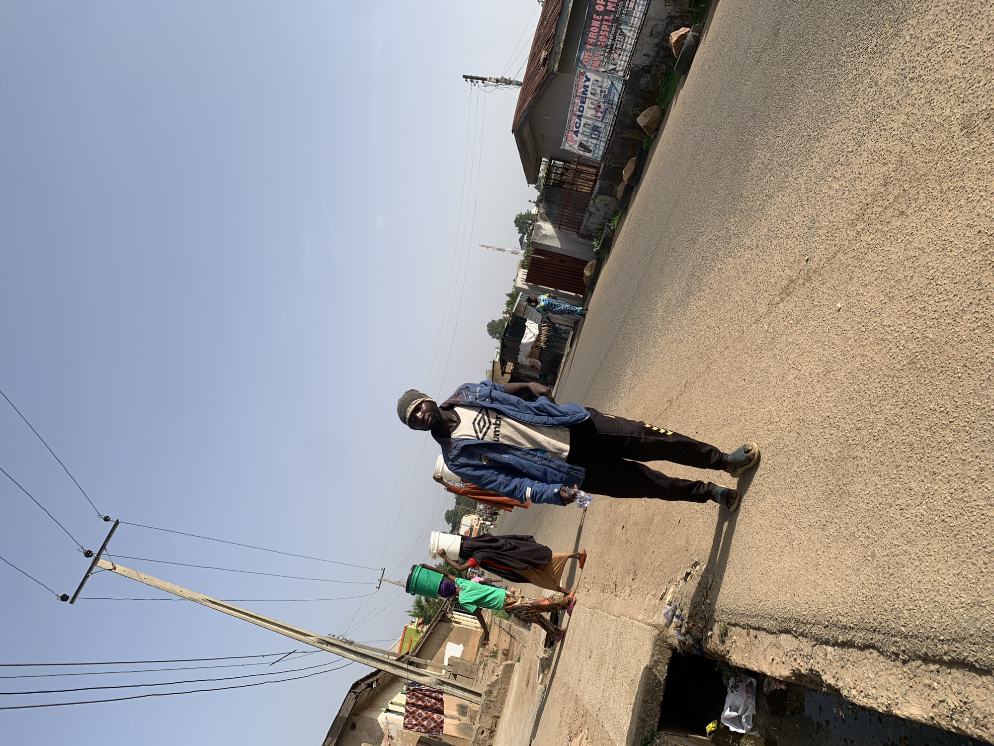 The image captures an outdoor scene in Jos, Nigeria, featuring a man in a blue denim jacket, a white t-shirt with a logo, black trousers, and a dark beanie. He stands on the edge of a paved road with a gutter running alongside it, holding a water bottle in his right hand. In the background, there are other individuals carrying buckets on their heads, suggesting a water collection activity. A utility pole with power lines is visible on the left side of the frame, while buildings and billboards can be seen on the right side of the road. The sky is light blue, indicating daylight.