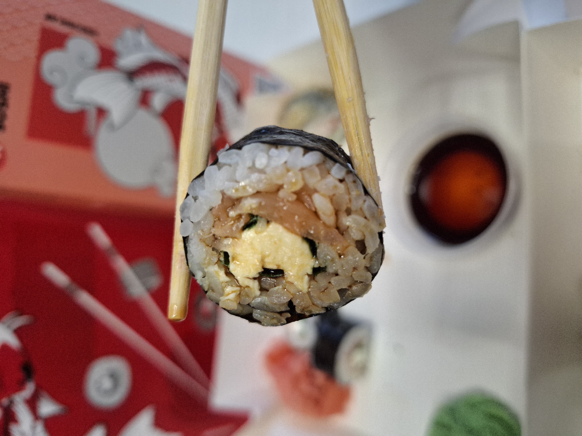 A close-up, eye-level, outdoor shot shows a single sushi roll held by two wooden chopsticks. The sushi roll is in focus and appears to be a maki roll with rice and seaweed. Inside the roll, there are visible fillings of scrambled egg and what looks like shredded salmon. The chopsticks are positioned horizontally, with the sushi roll suspended between them. In the background, out of focus, is a bright red container with white graphics, possibly a takeout box. To the left and below the sushi roll, there are other out-of-focus elements that resemble a dollop of green wasabi and a small dish of soy sauce. The overall lighting suggests it is daytime. The scene does not contain any identifiable landmarks or elements that would suggest a specific location in Bratislava, Slovakia.